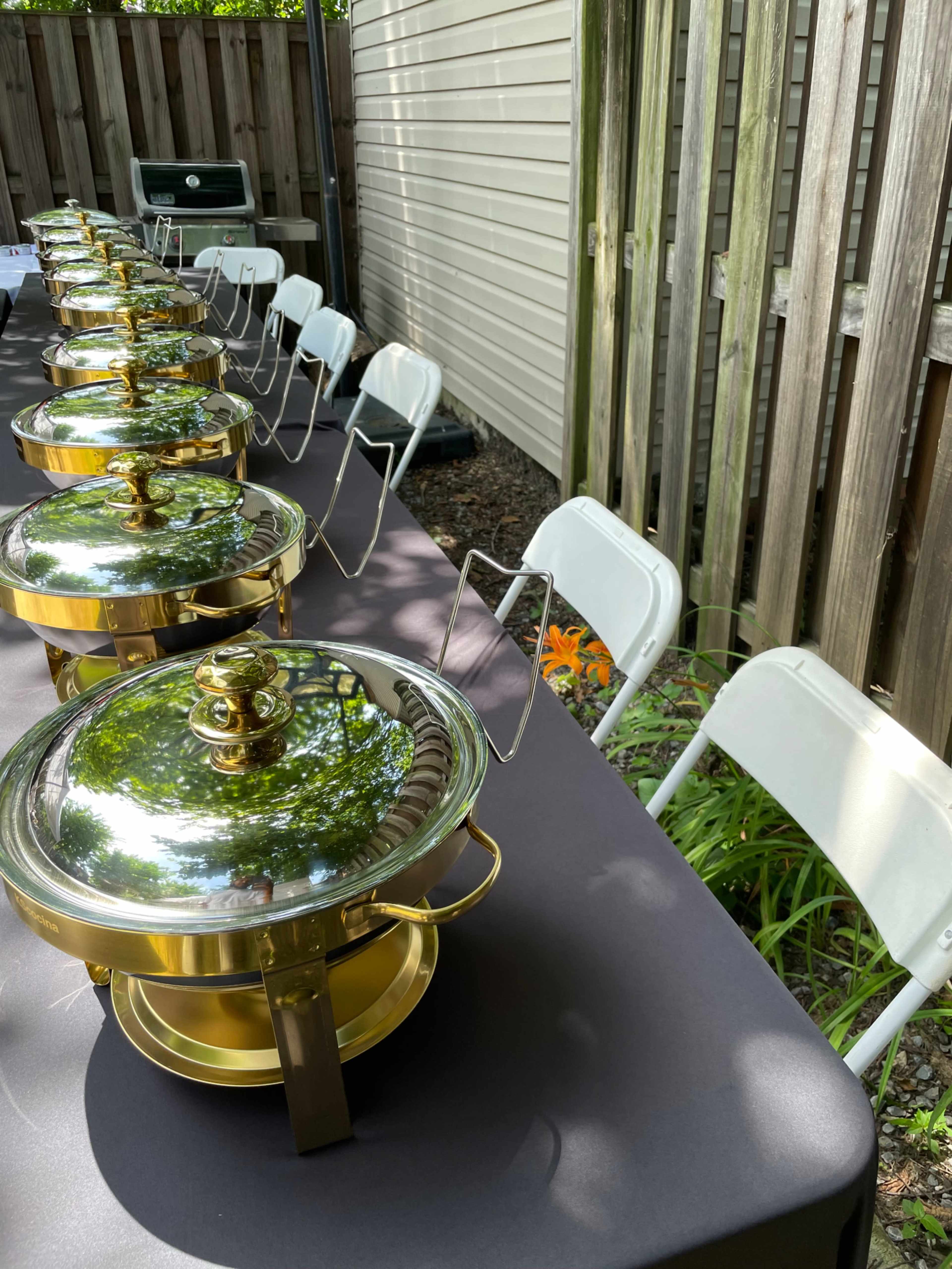 A row of brass chafing dishes is set on a table outside, near a wooden fence and surrounded by greenery.