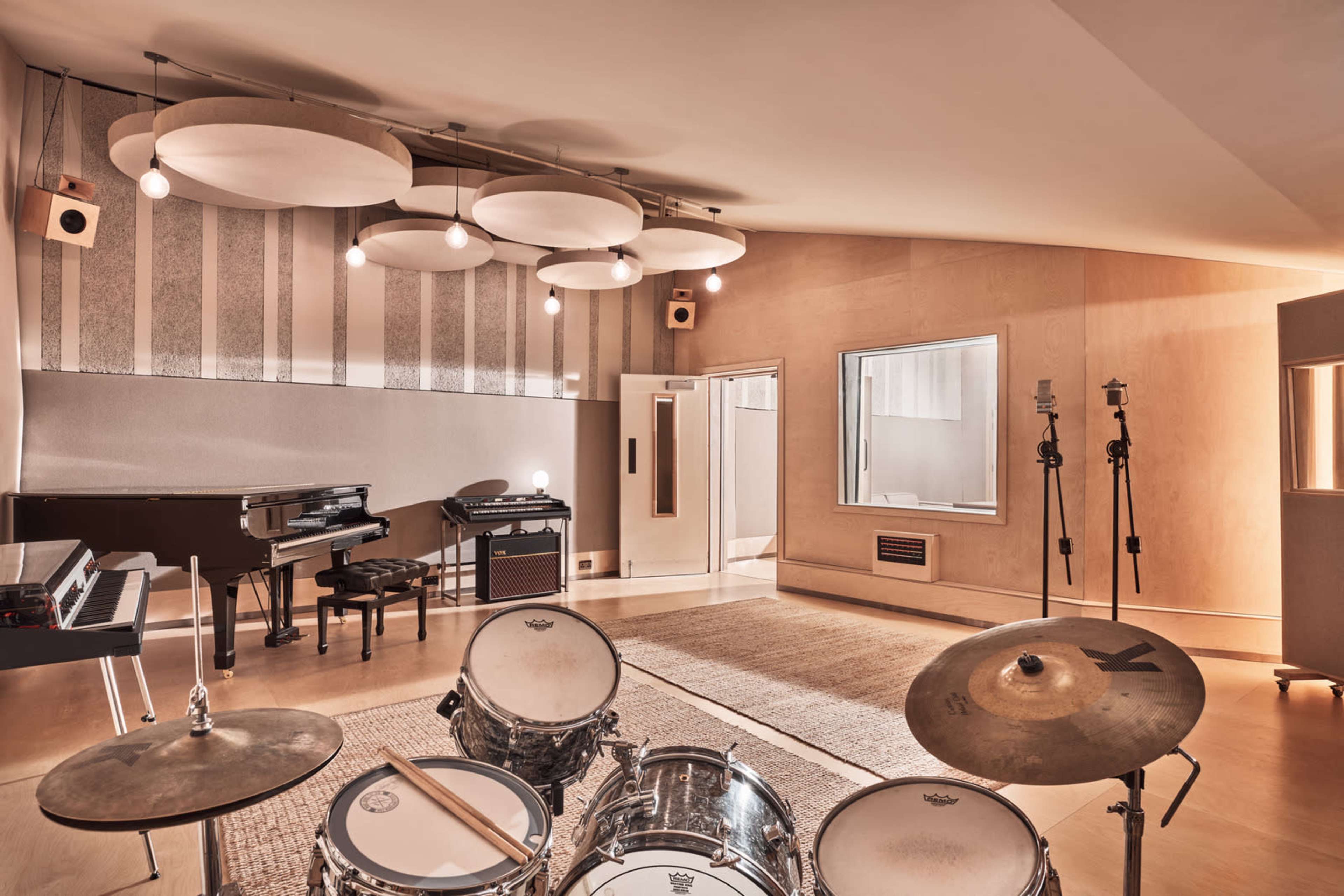 A spacious music studio features a drum set in the foreground, a piano, and several soundproofing panels, with prominent circular light fixtures overhead.