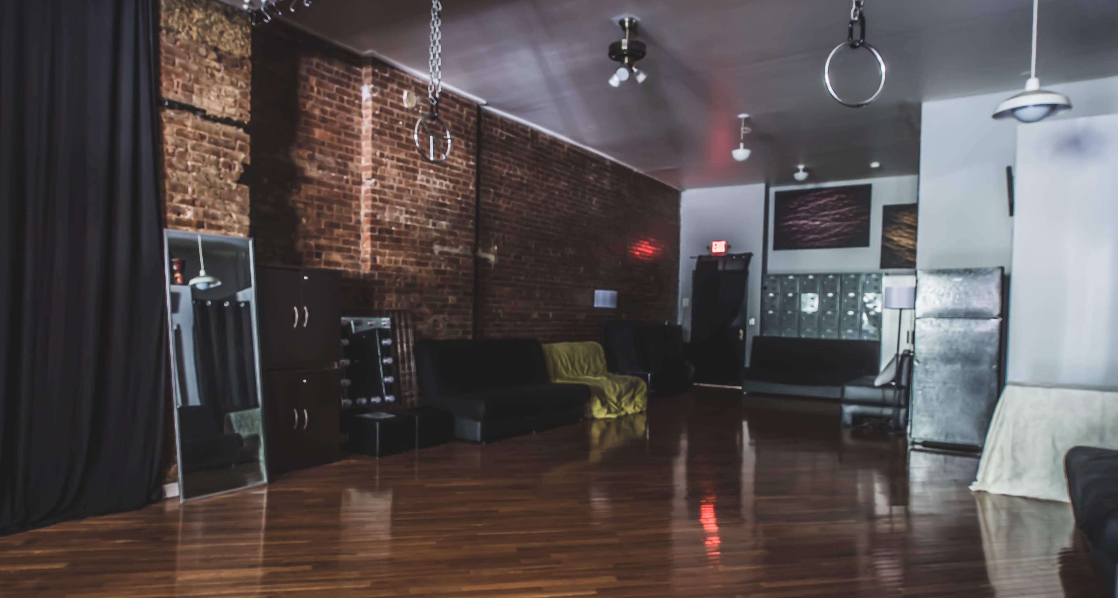 The image shows a spacious interior with wooden floors, exposed brick walls, mirrors, and a few pieces of black furniture, illuminated by overhead lighting.