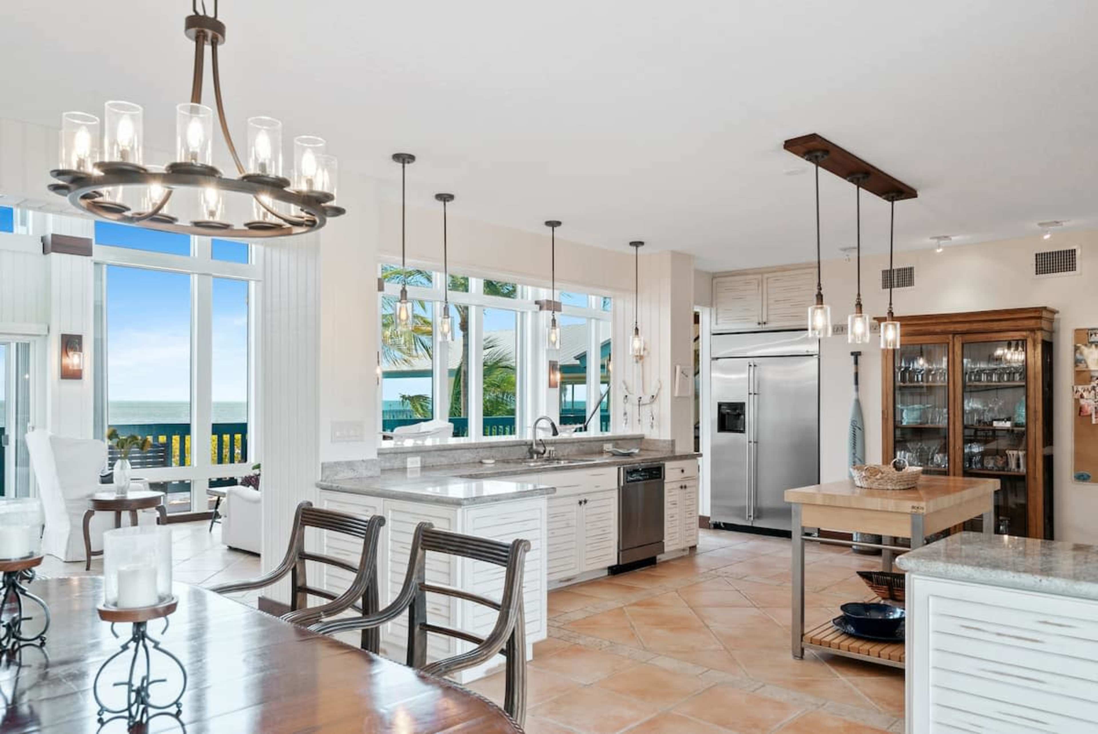 The image shows a modern kitchen with a large island, stainless steel appliances, and windows offering a view of the ocean.
