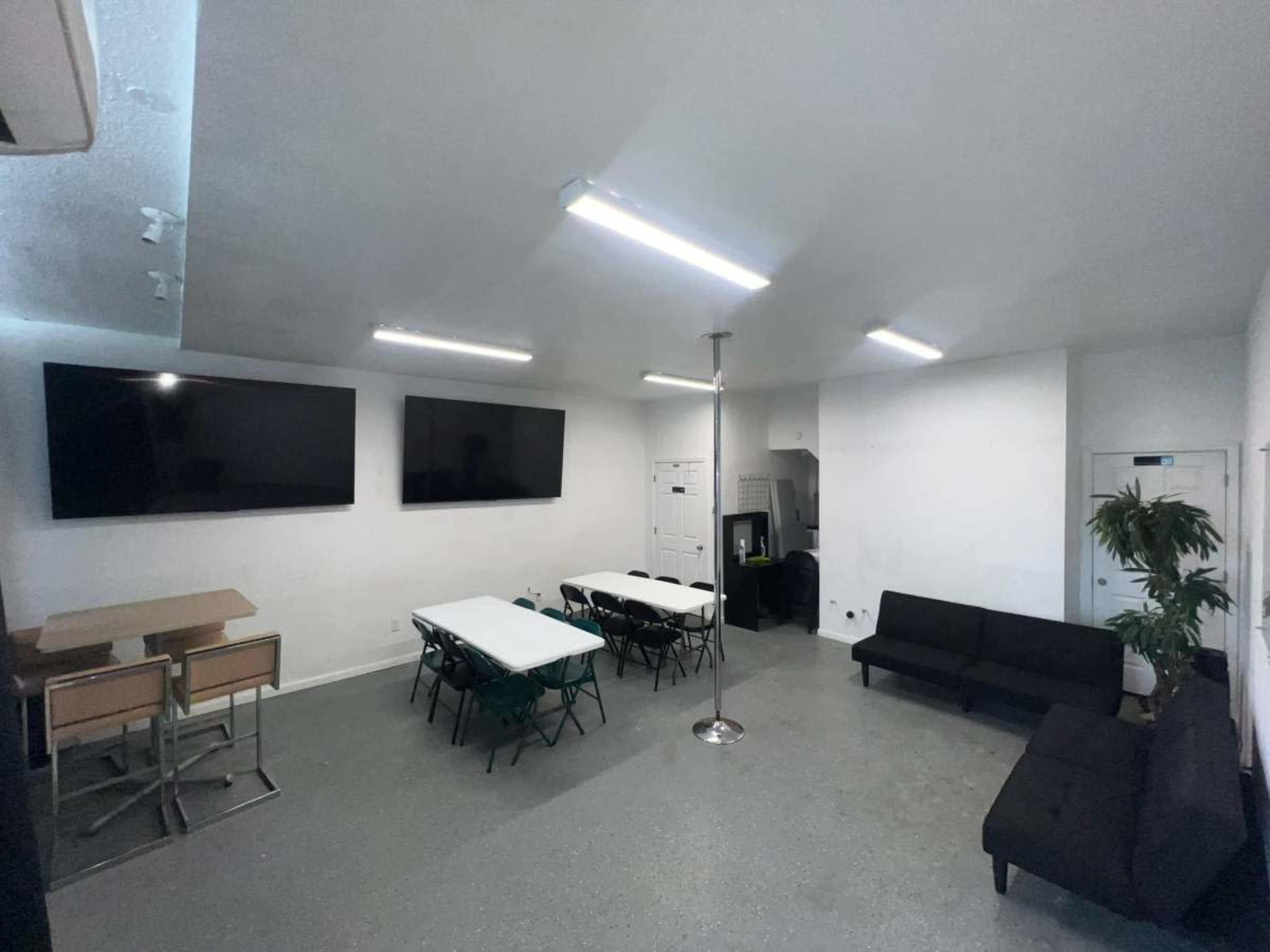 The image shows a spacious room with two large television screens on the wall, a table with chairs, a black couch, and a pole in the center.
