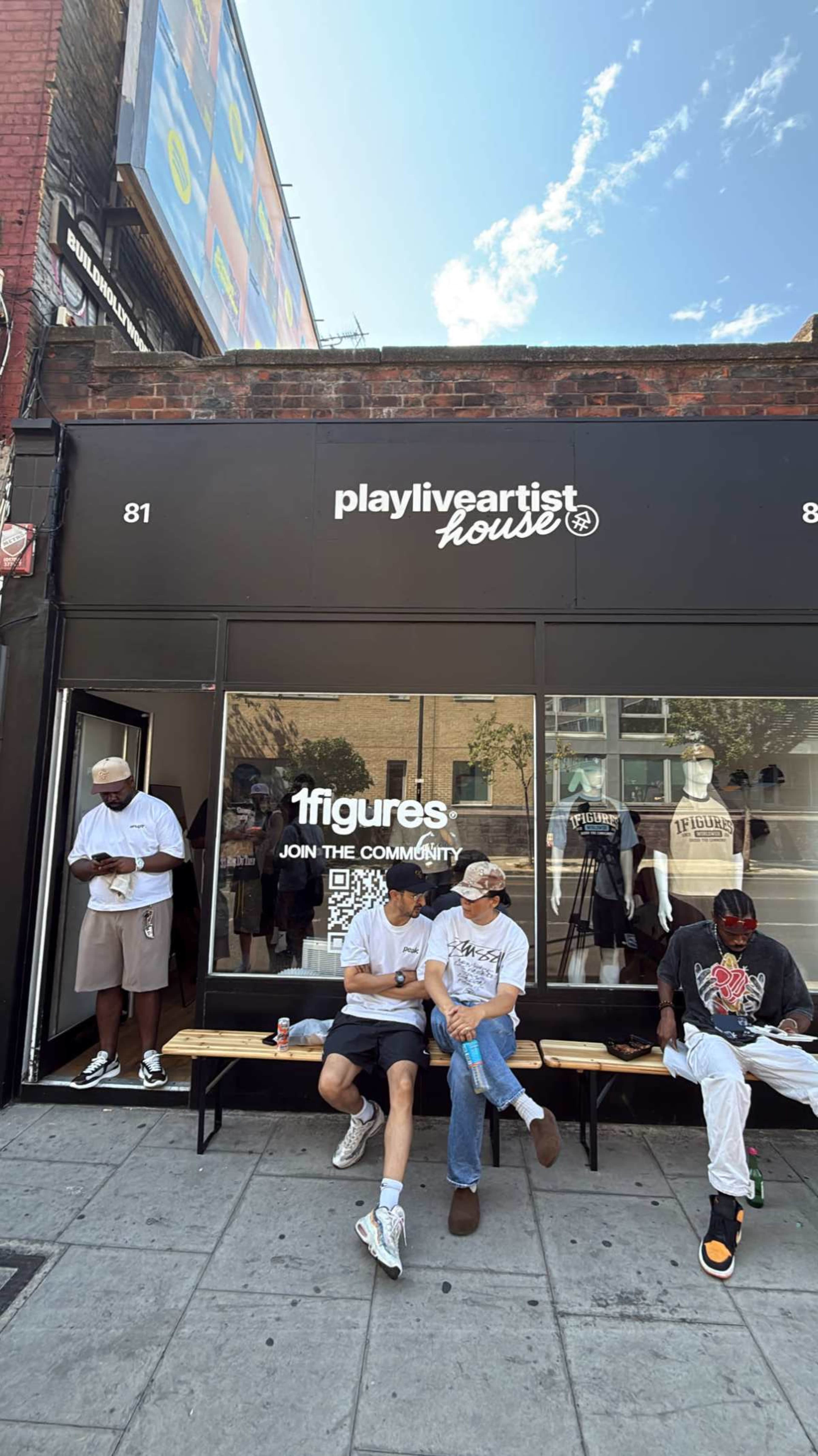 The image shows a storefront labeled "playliveartisthouse," with three individuals sitting on a bench outside and a fourth person standing nearby, all in urban clothing.