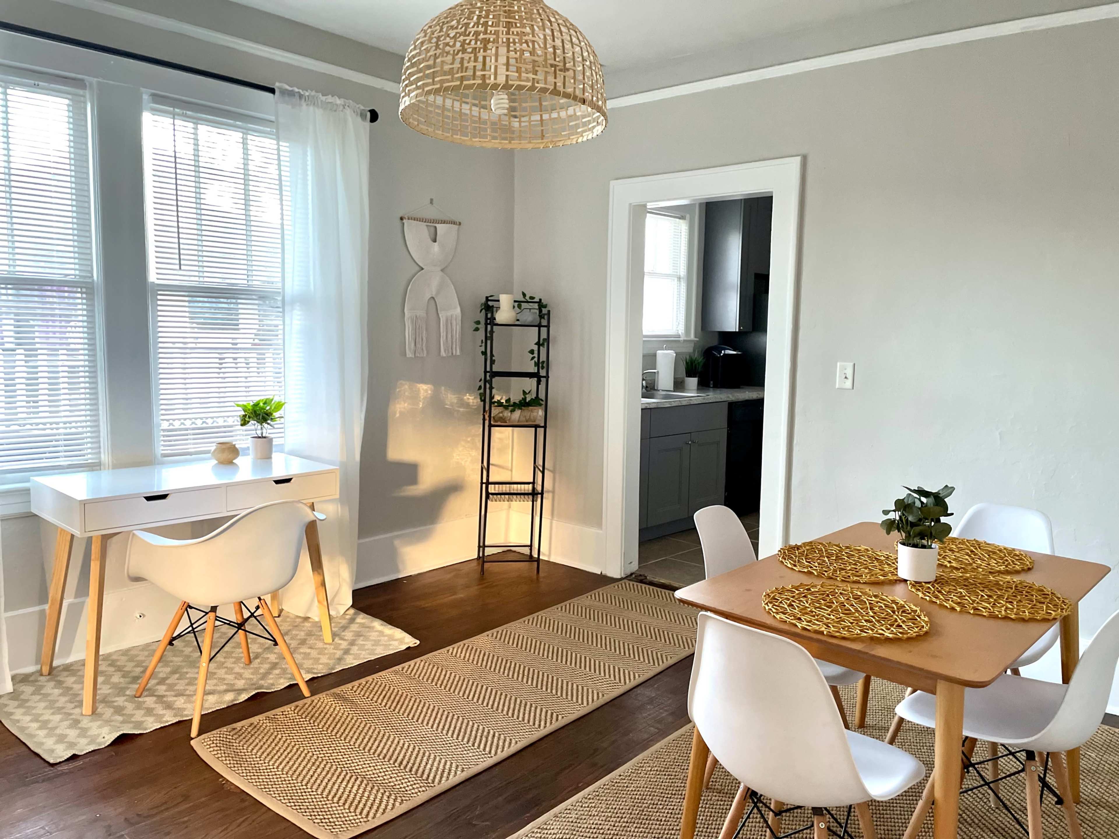 A bright dining area features a wooden table with white chairs, a desk with a plant, and a woven light fixture hanging from the ceiling.