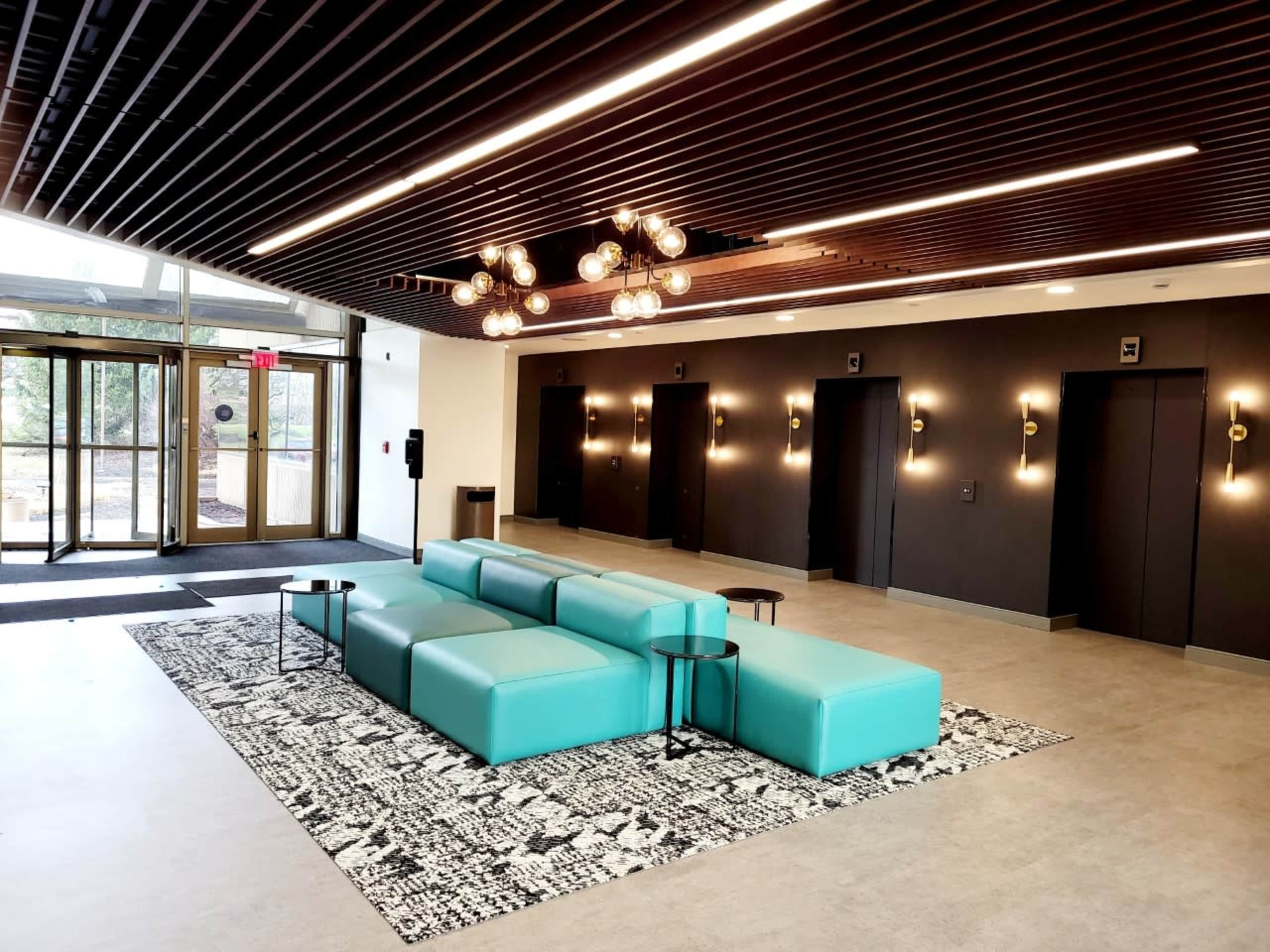 The image shows a modern lobby with a turquoise sectional sofa, a patterned rug, and elevators along the walls.
