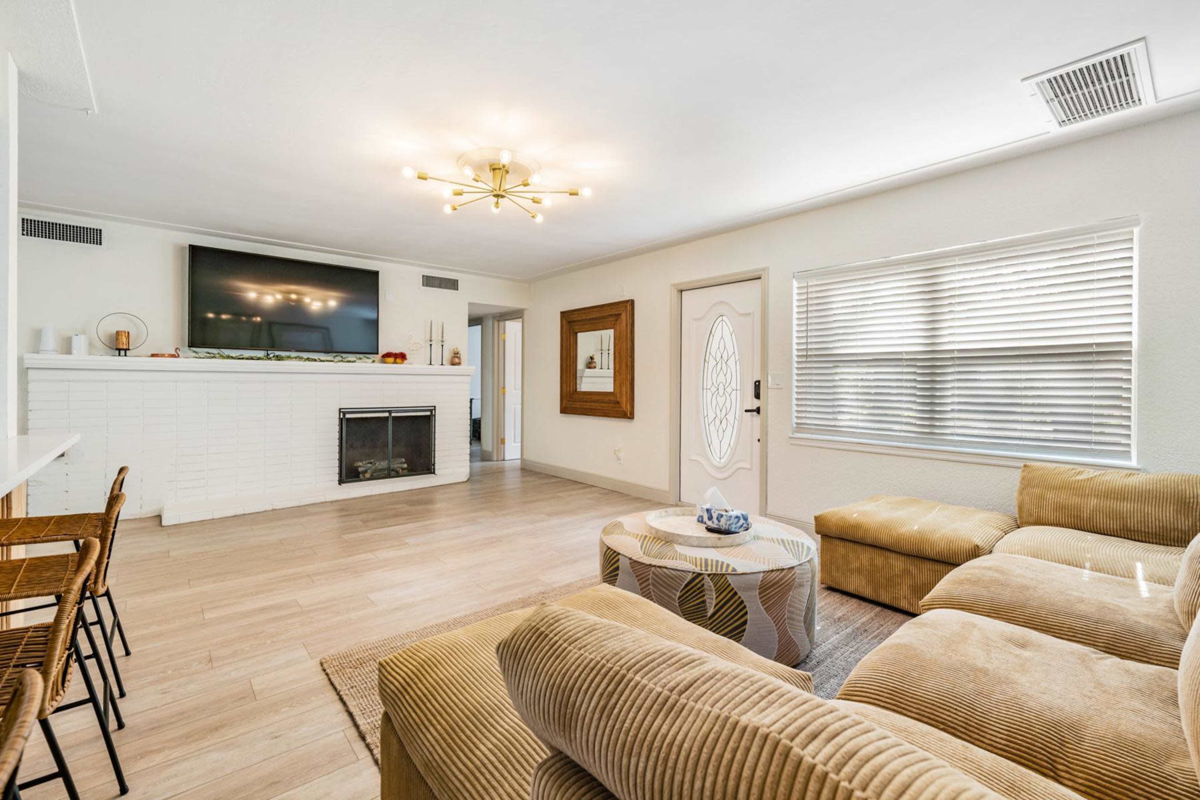 The living room features a sectional sofa, a circular coffee table, a flat-screen TV mounted above a white brick fireplace, and a front door with a decorative window.