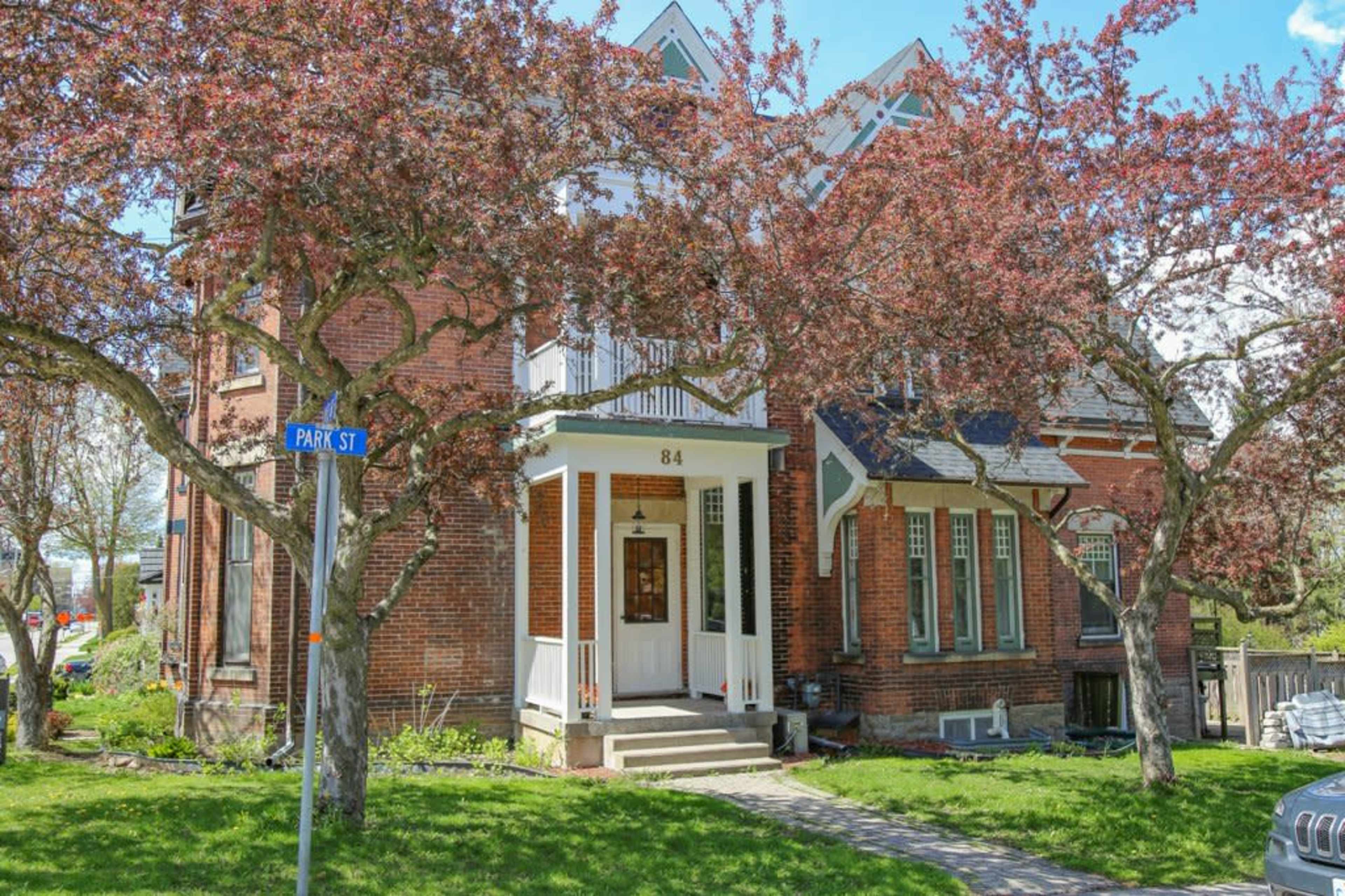 A brick house with a white front door and large windows is surrounded by blooming trees and a street sign indicating "Park St."