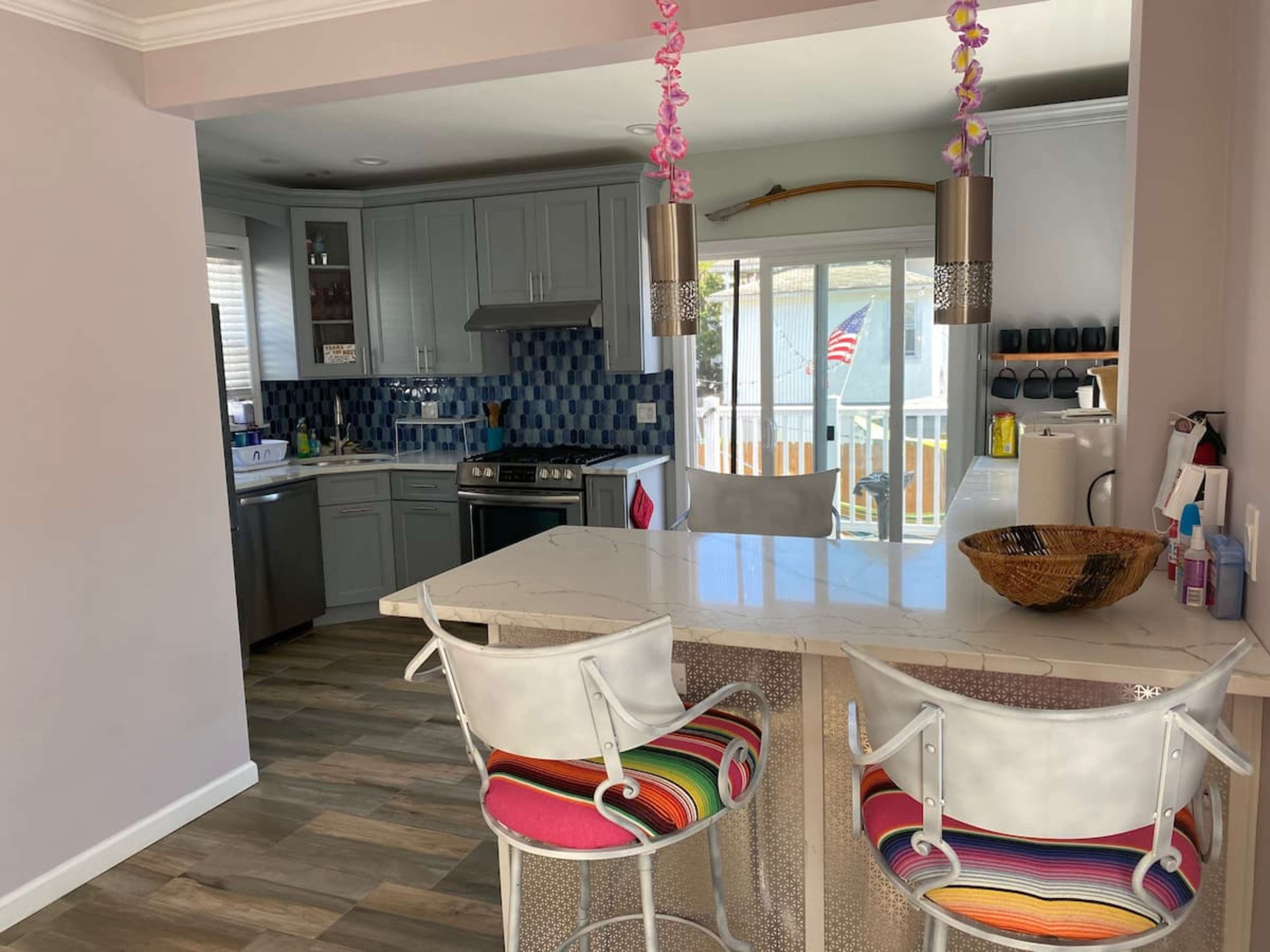 A modern kitchen features gray cabinets, a countertop with colorful bar stools, and a view of a deck with an American flag in the background.