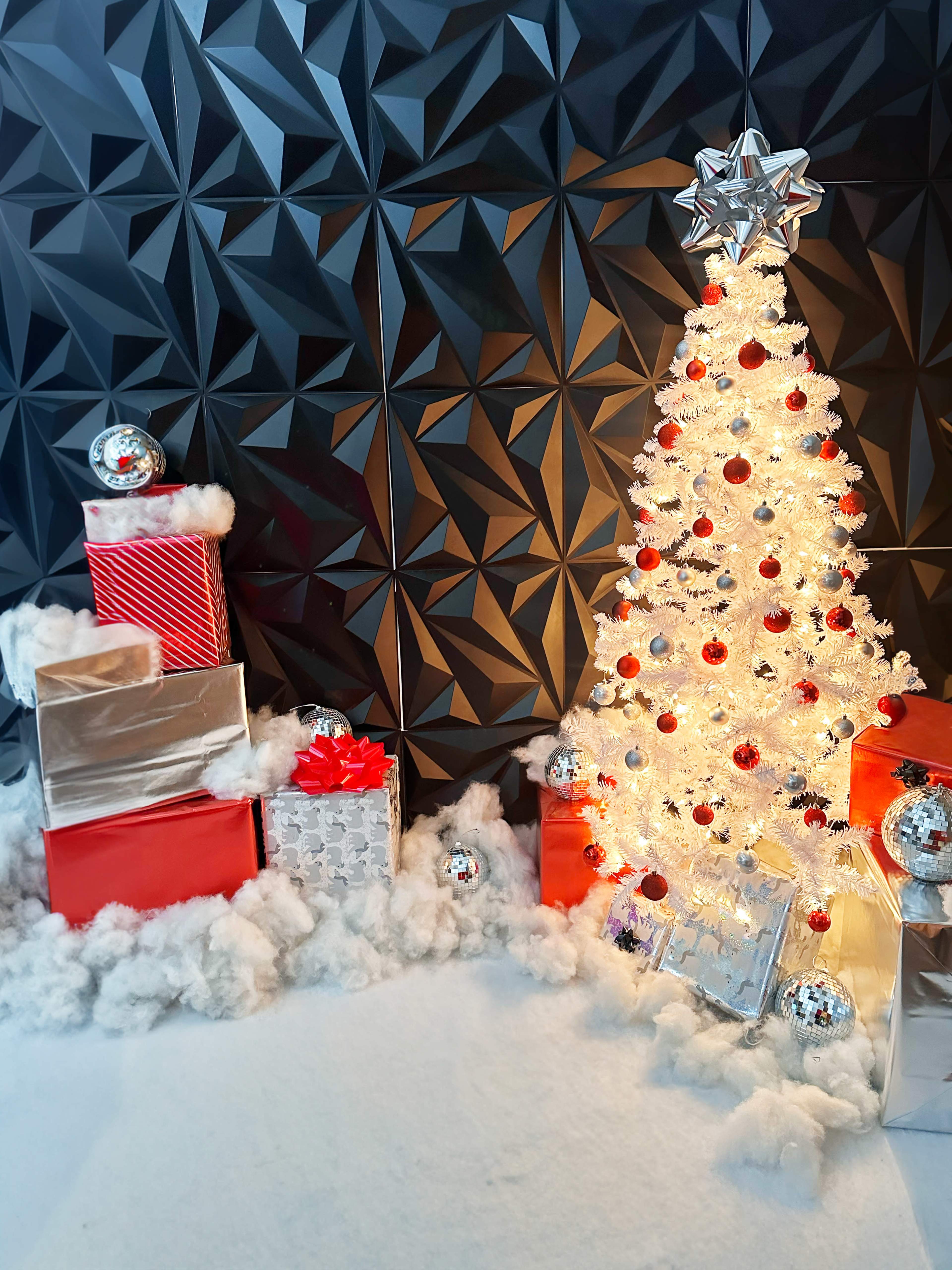 The image shows a decorated Christmas tree with white lights and ornaments next to neatly arranged gift boxes in a snowy setting against a textured black wall.