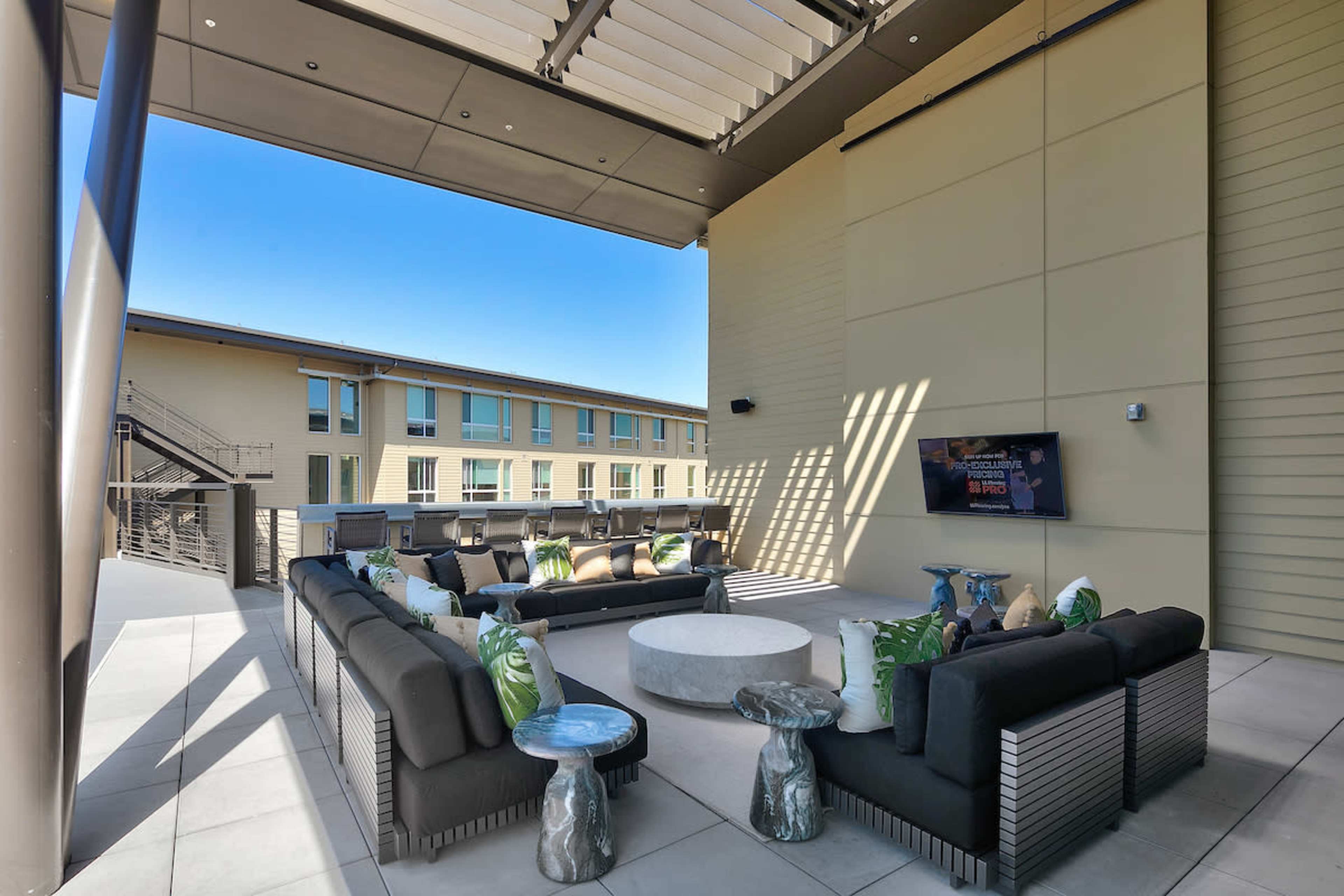 A modern outdoor lounge area featuring sofas arranged around a circular table, with an overhead covering and a television mounted on the wall.