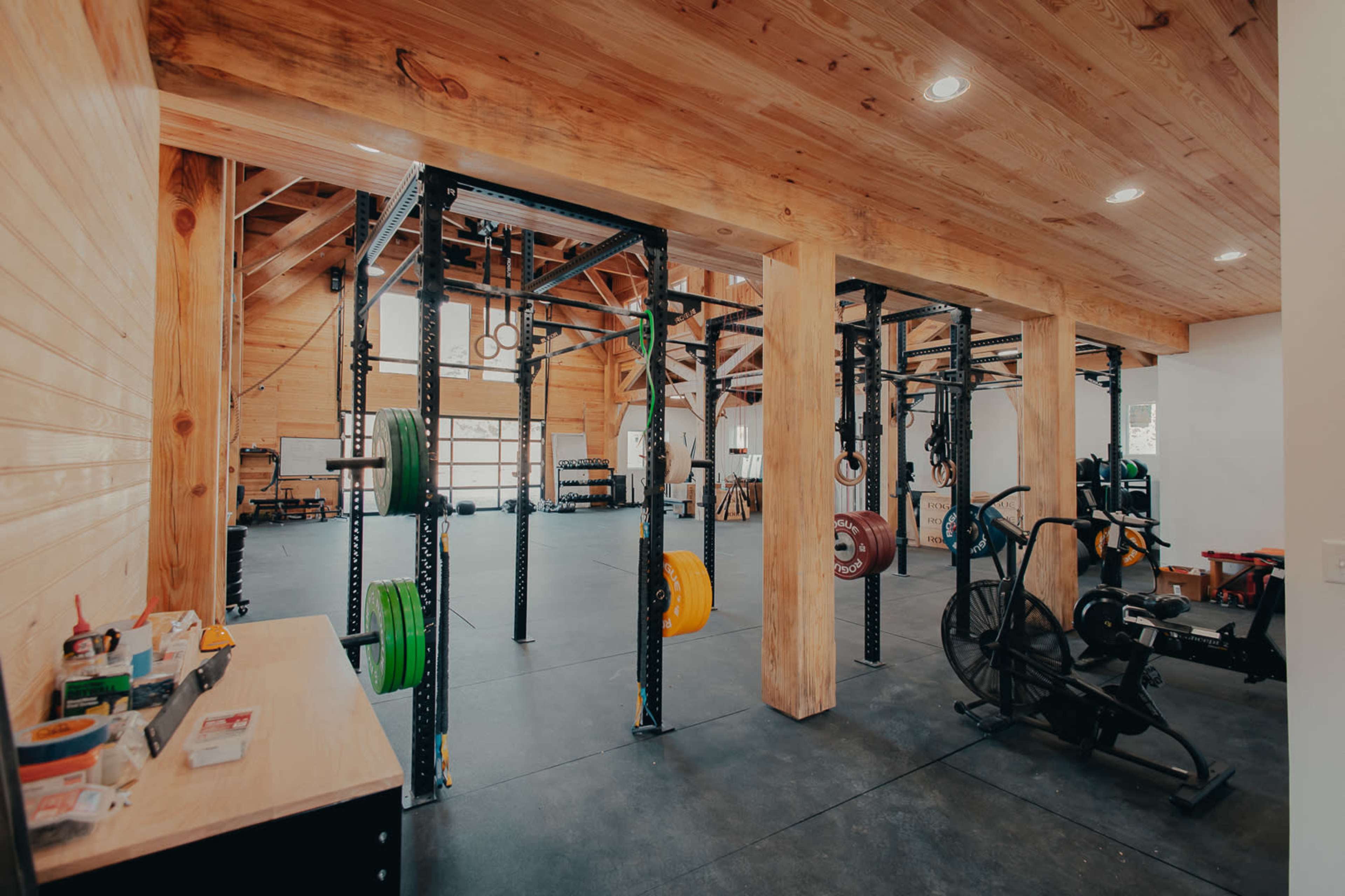 The image shows a well-equipped gym featuring weightlifting racks, various fitness equipment, and a spacious wooden interior.