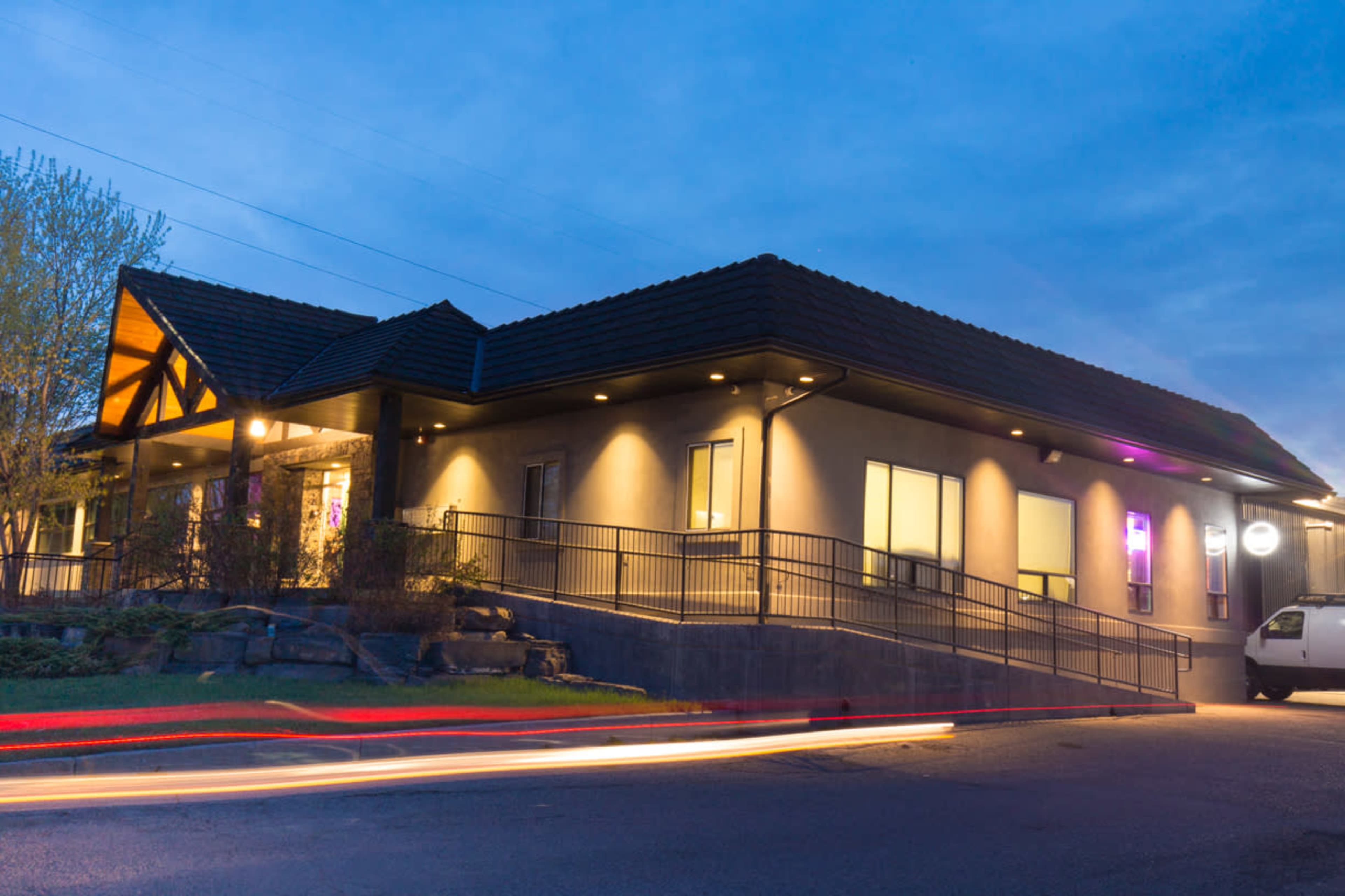 The image shows a modern building with a sloped roof, illuminated windows, and a ramp leading to the entrance, set against a twilight sky.