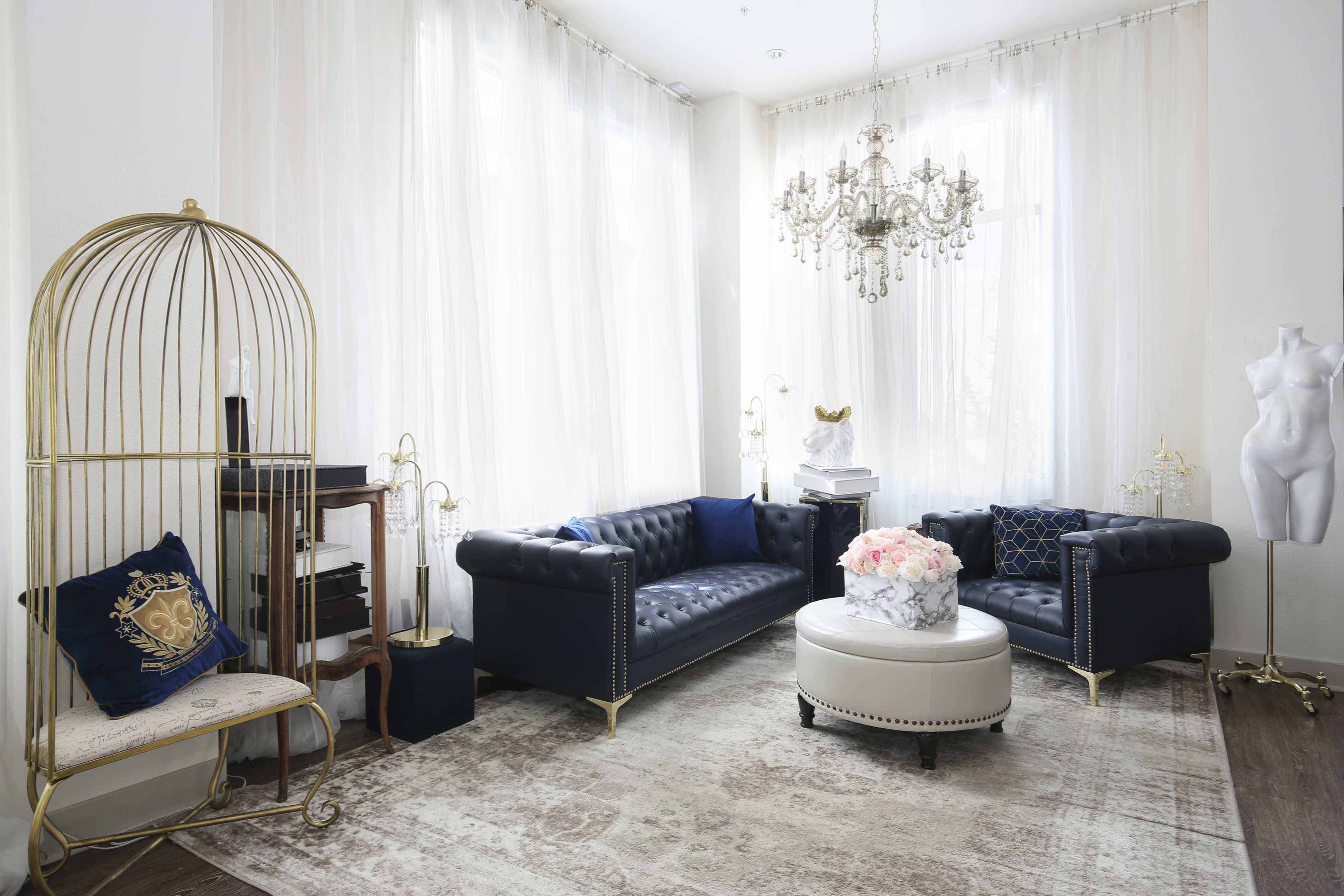 The image features a stylish living room with two black chesterfield sofas, a round white coffee table, a decorative chandelier, and a mannequin, all set against light, sheer curtains.