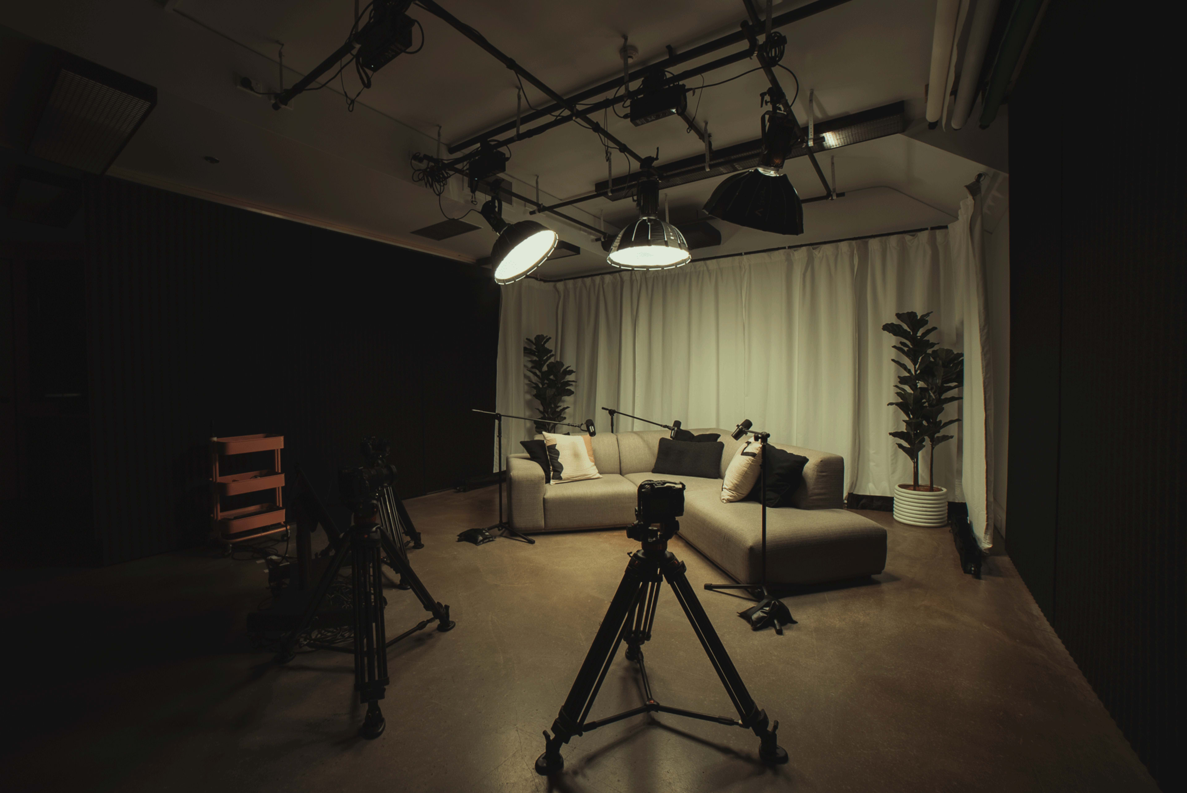 The image shows a dimly lit studio with two sofas, microphone stands, and cameras positioned around a backdrop of curtains and potted plants.