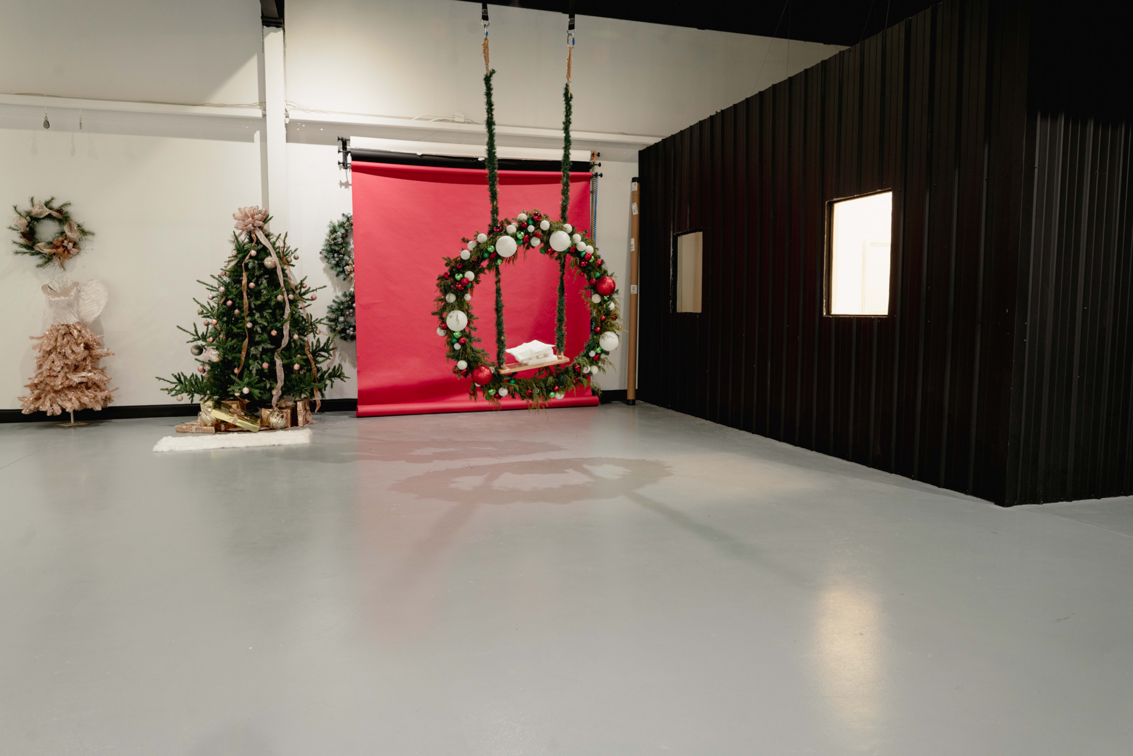 The image shows an empty studio space with a red backdrop, a decorative Christmas wreath hanging from the ceiling, and a Christmas tree alongside a mannequin dressed in festive attire.