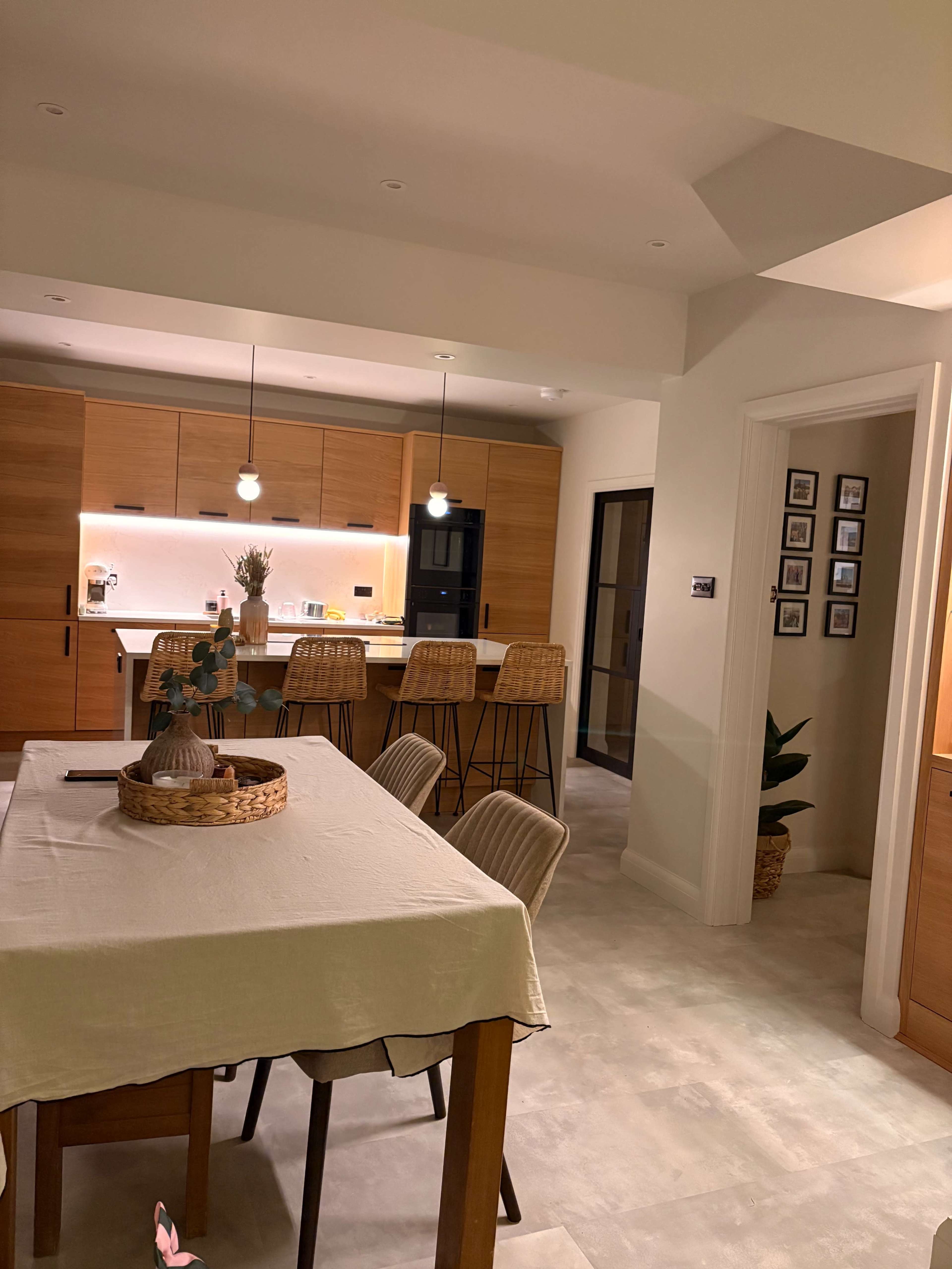 The image depicts a modern kitchen and dining area featuring wooden cabinetry, a dining table with a tablecloth, and bar stools at a kitchen island.