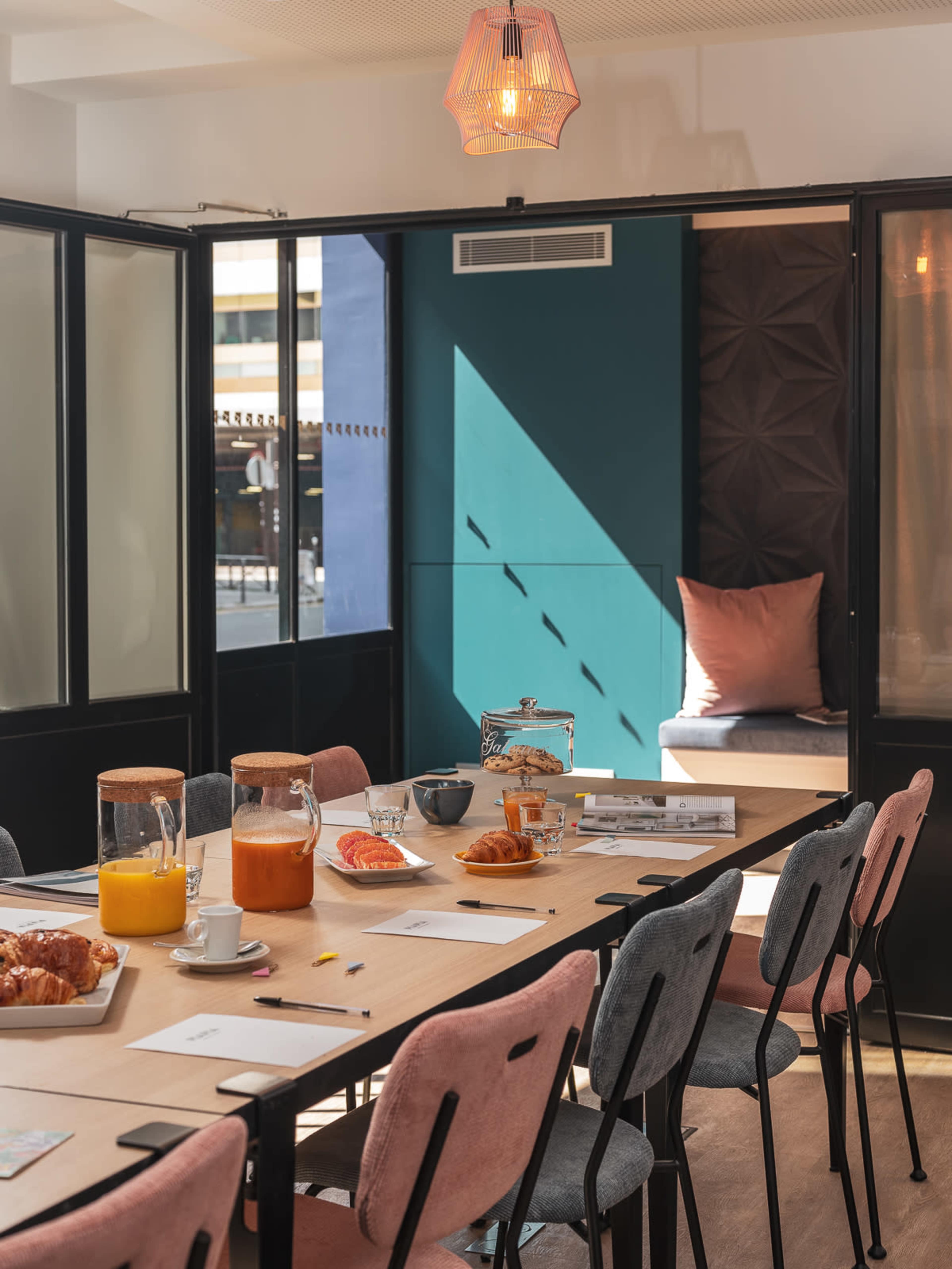 A meeting room features a long table set with drinks, pastries, and stationery, illuminated by natural light streaming through large windows.