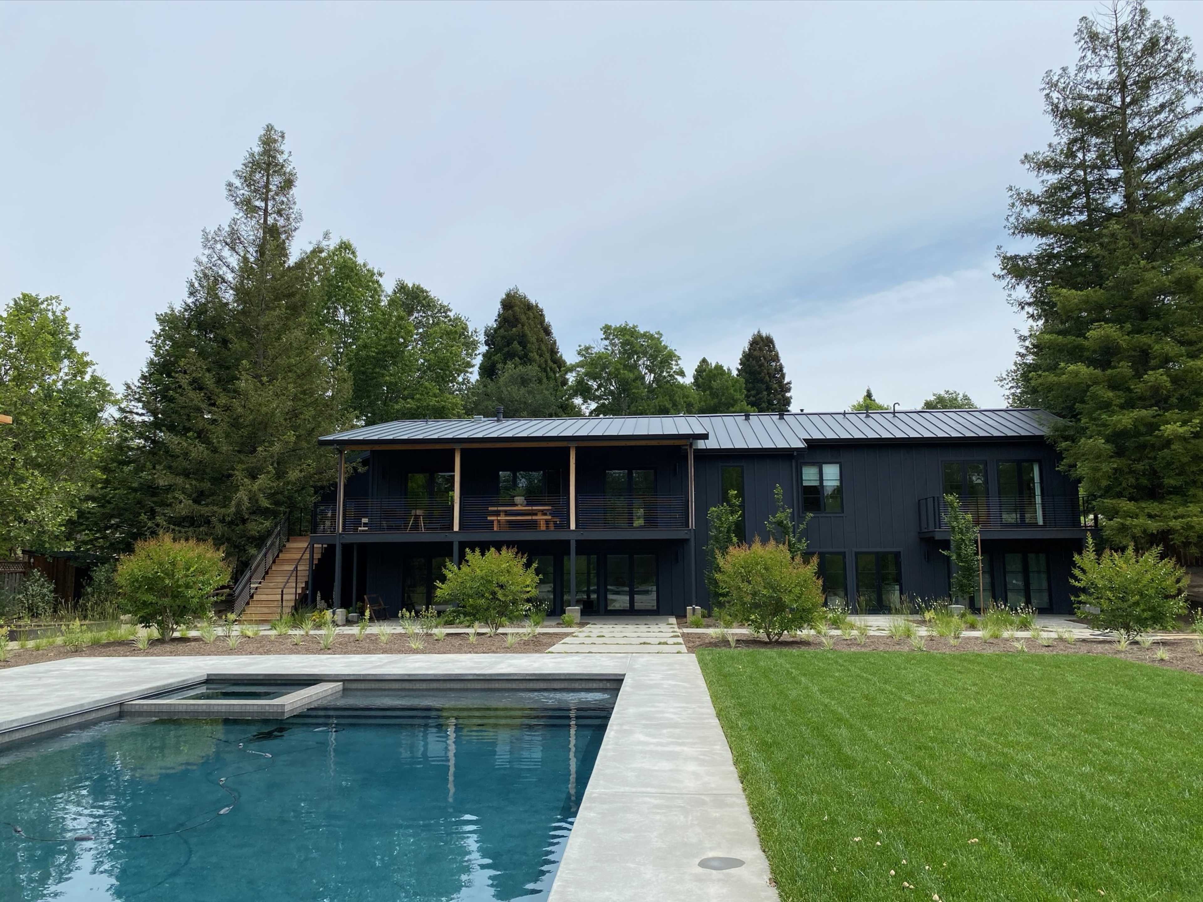 A modern two-story black house with a metal roof overlooks a swimming pool surrounded by greenery and landscaping.