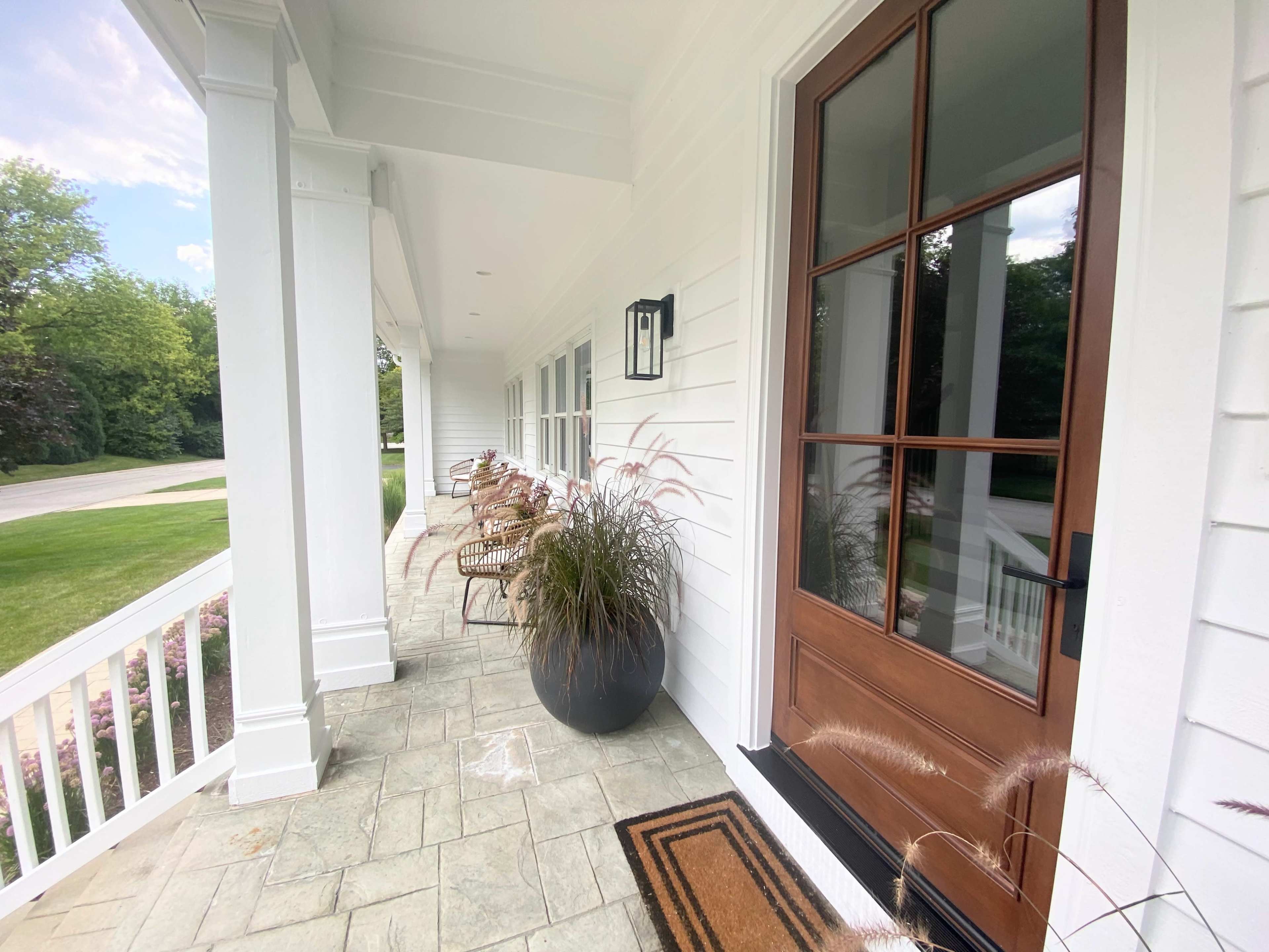 The image shows a welcoming porch with a wooden front door, a decorative mat, and modern seating arranged along the side.
