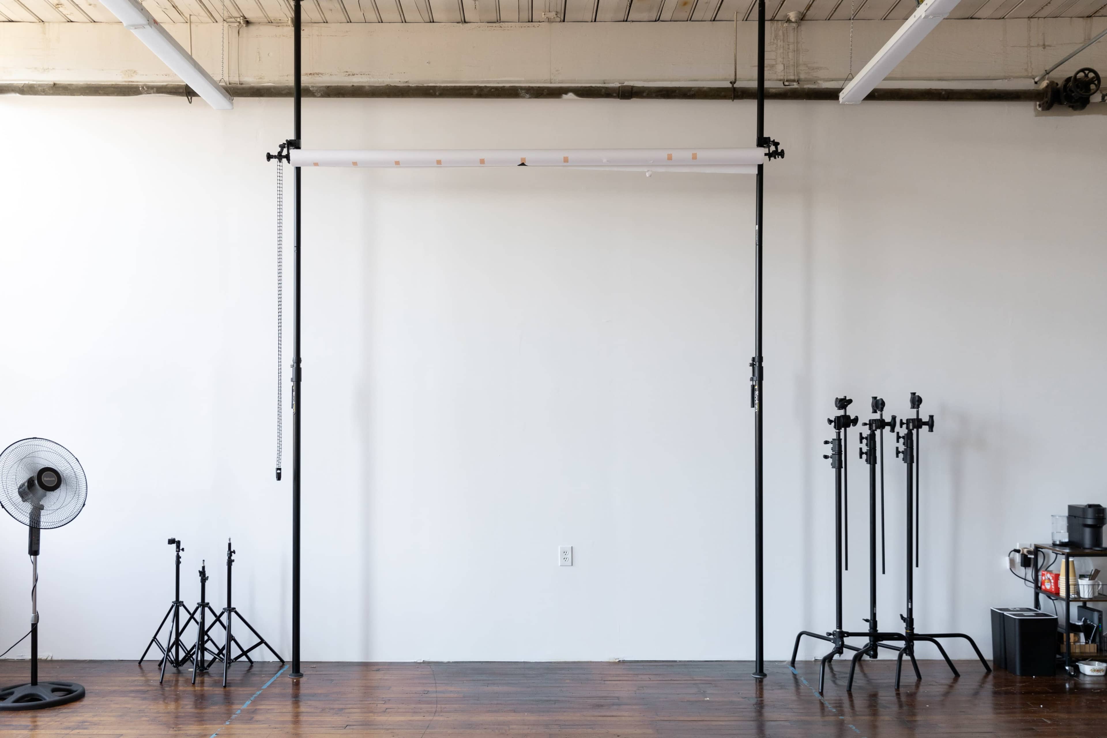 A minimalistic studio space features a backdrop support system, various light stands, a fan, and a coffee maker against a plain wall.