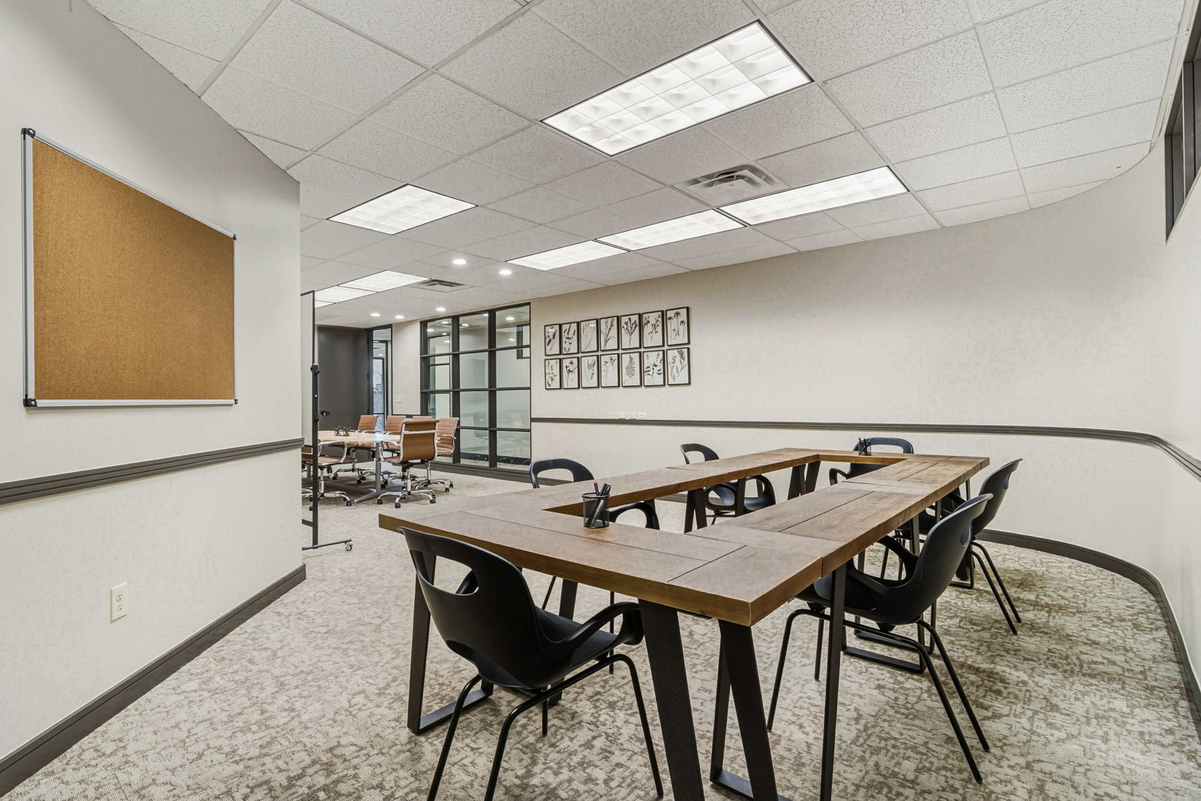 The image shows a conference room with a long wooden table surrounded by black chairs, and a bulletin board on the wall.