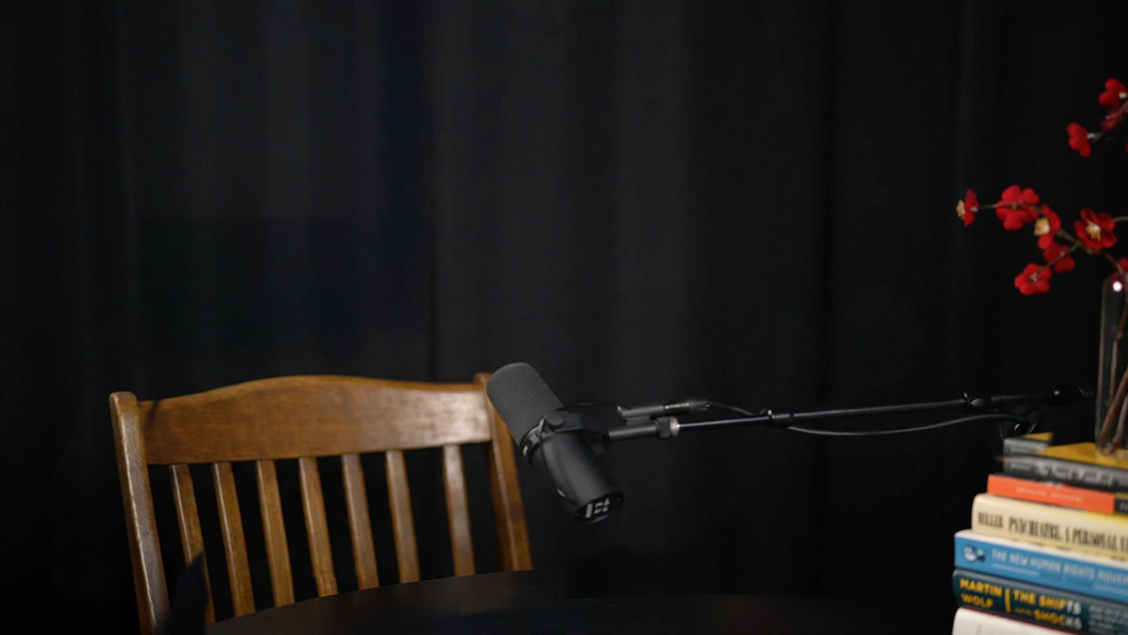 The image shows a wooden chair next to a microphone on a stand, with a black backdrop and a stack of books on the table.