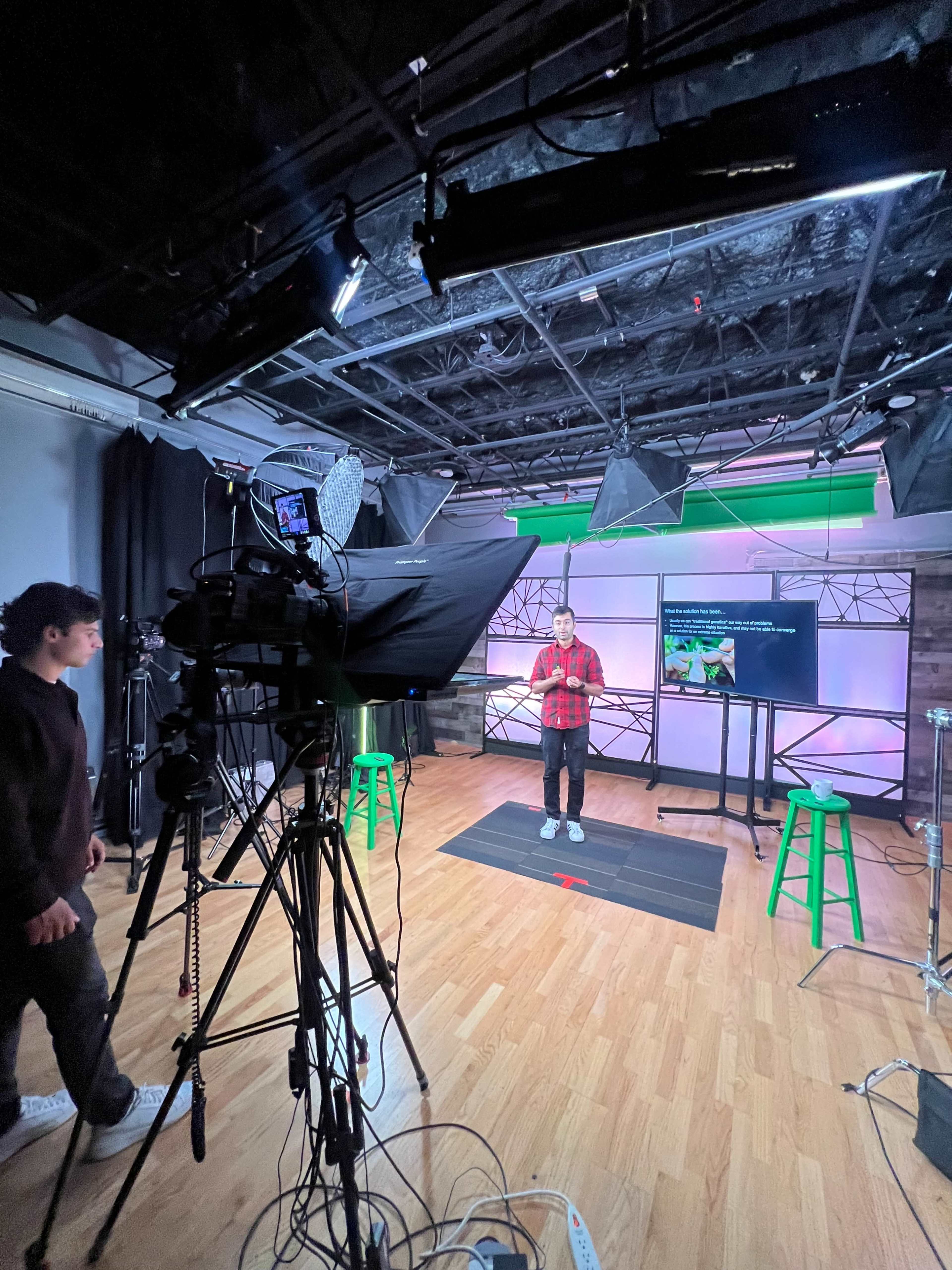 A presenter stands in a television studio, speaking into the camera while a crew member adjusts equipment in the background.