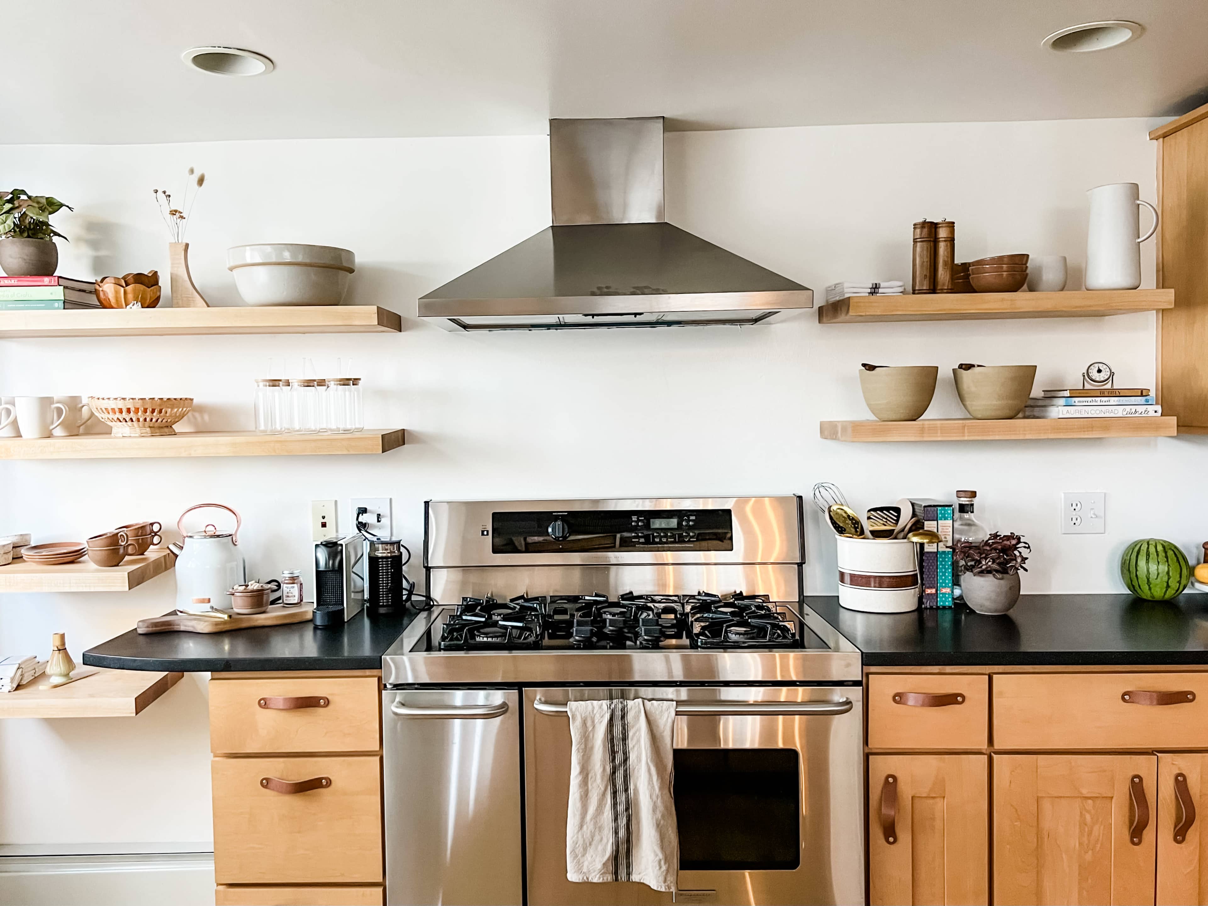 A modern kitchen with a stainless steel stove, a metallic range hood, and wooden shelves displaying various kitchen items.