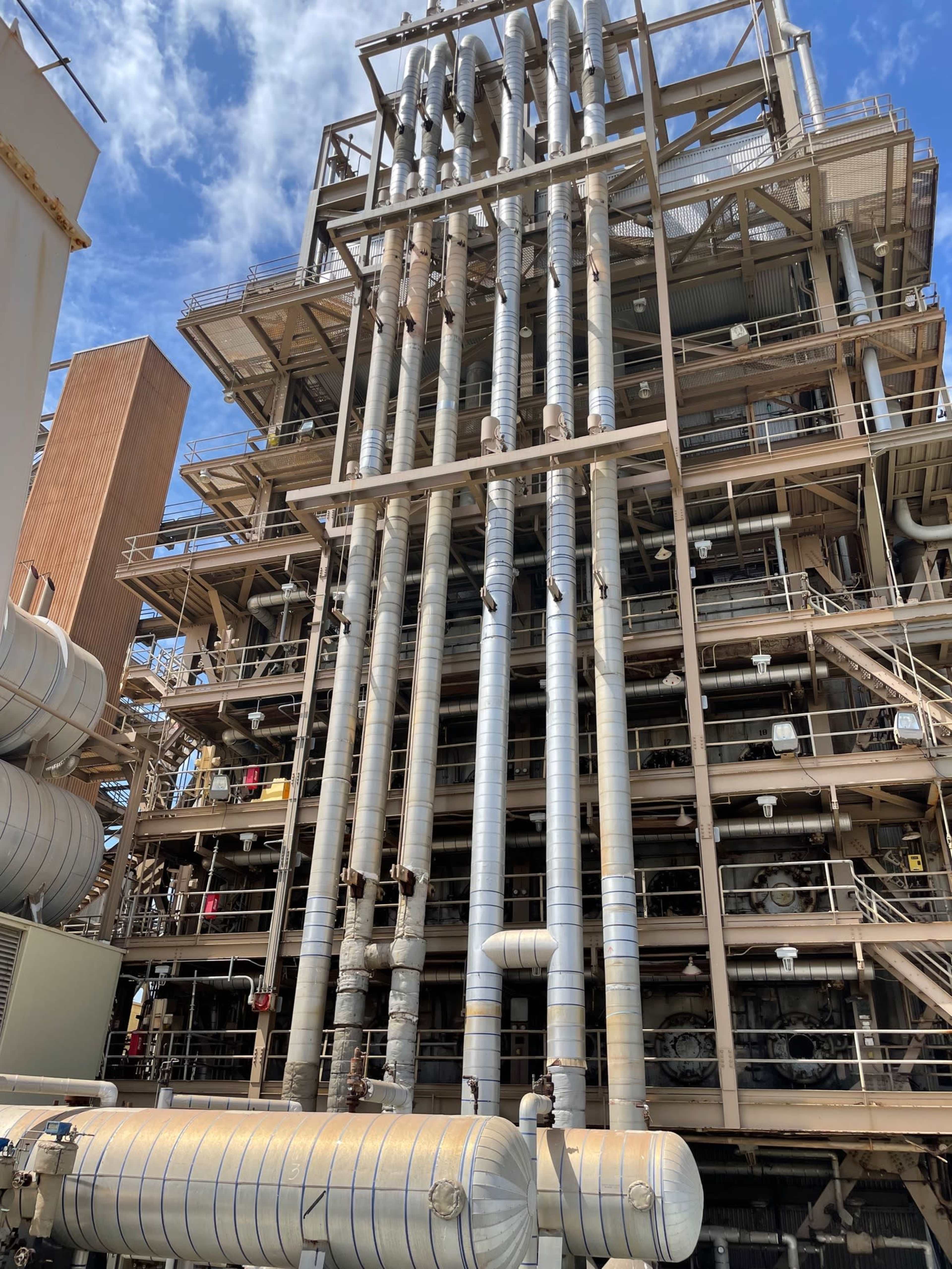 A large industrial structure featuring multiple vertical pipes and horizontal tanks against a blue sky.