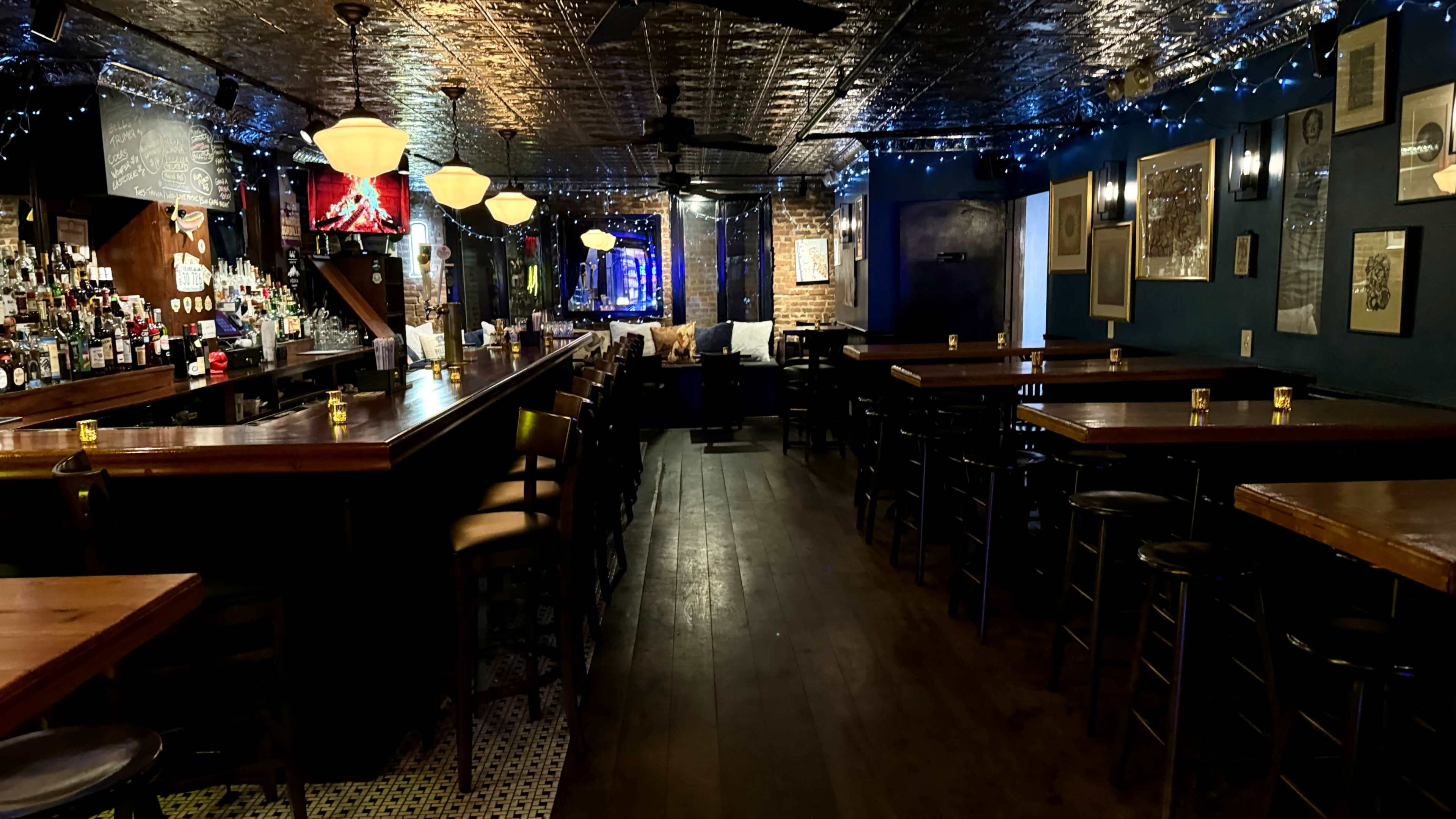 A dimly lit bar with a long wooden counter, stools, and scattered tables, adorned with string lights and artwork on the walls.