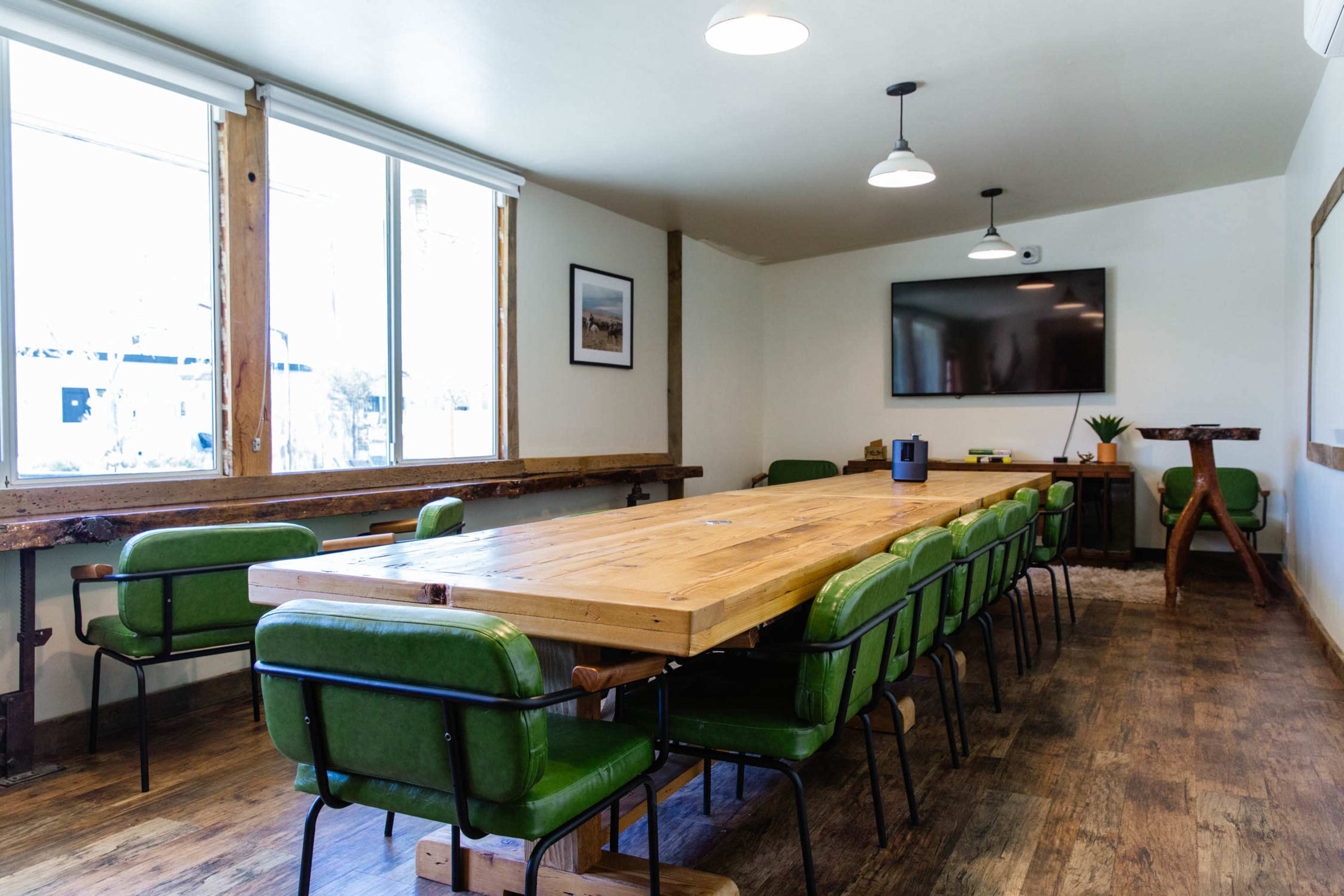 A modern conference room with a long wooden table surrounded by green chairs, large windows, and a wall-mounted television.