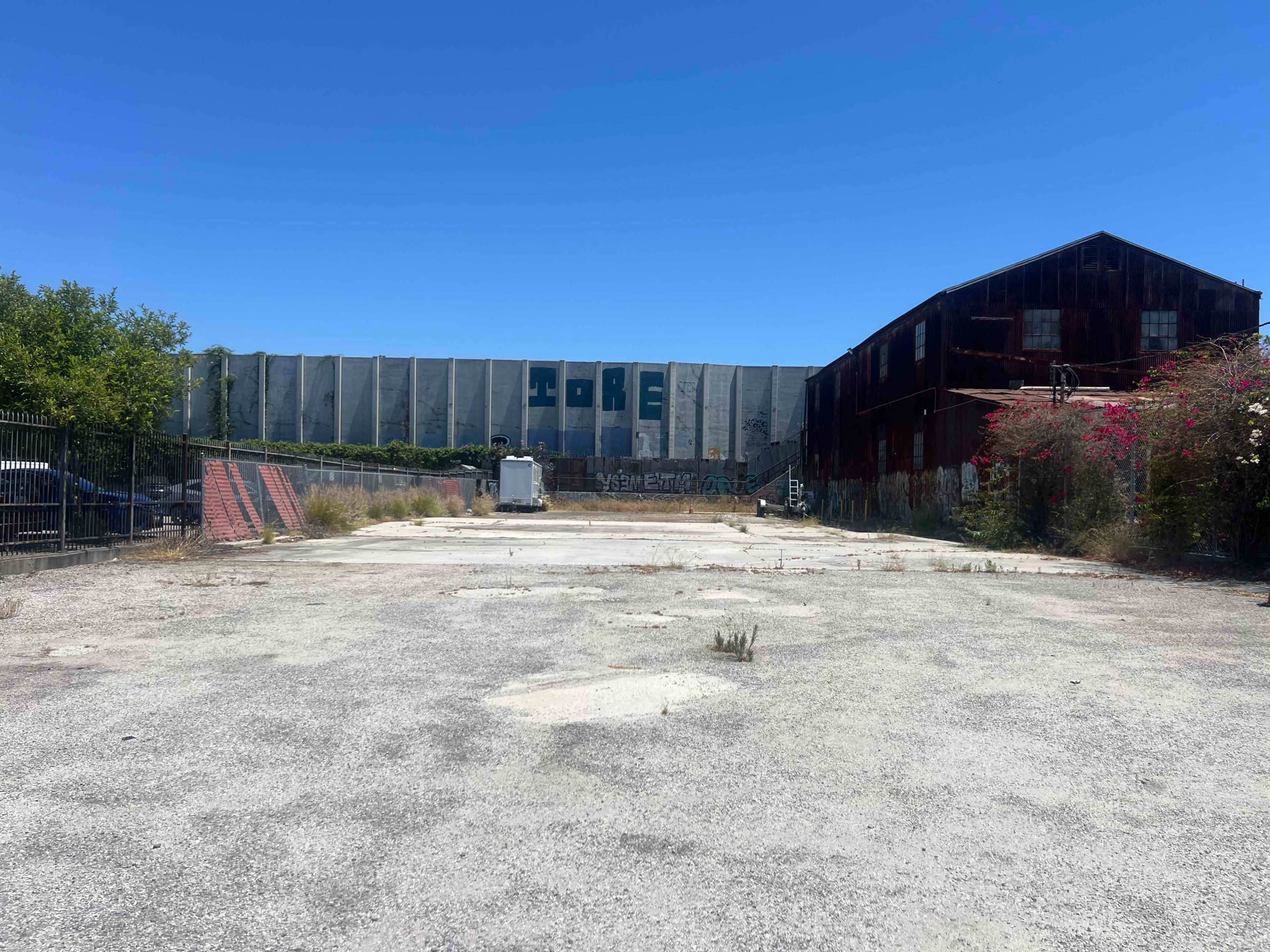 The scene shows an empty, gravel-covered lot with two large, dilapidated industrial buildings in the background under a clear blue sky.