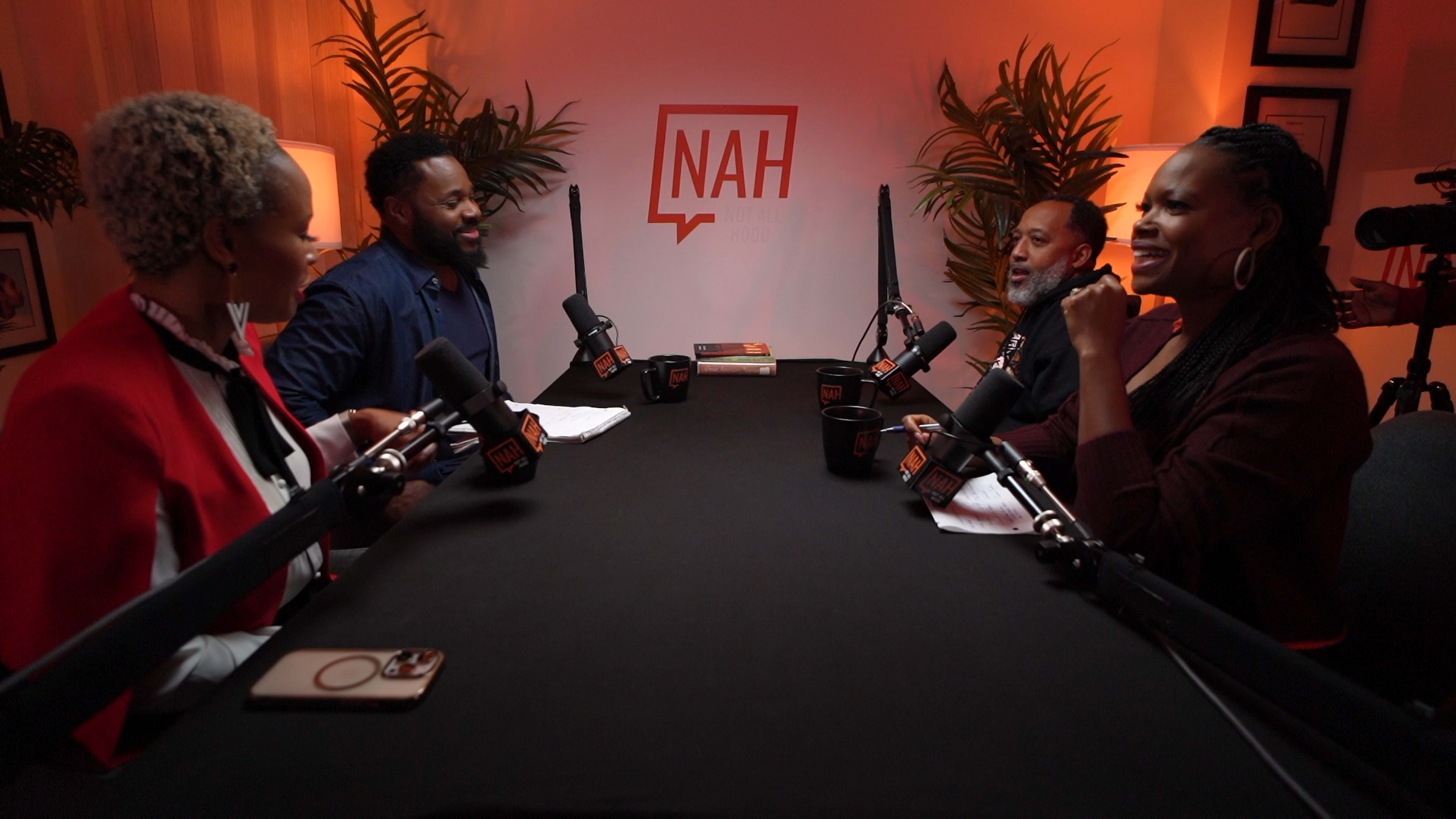 Four people engage in a discussion around a table with microphones, set in a brightly lit studio featuring plants and a backdrop with the word "NAH."