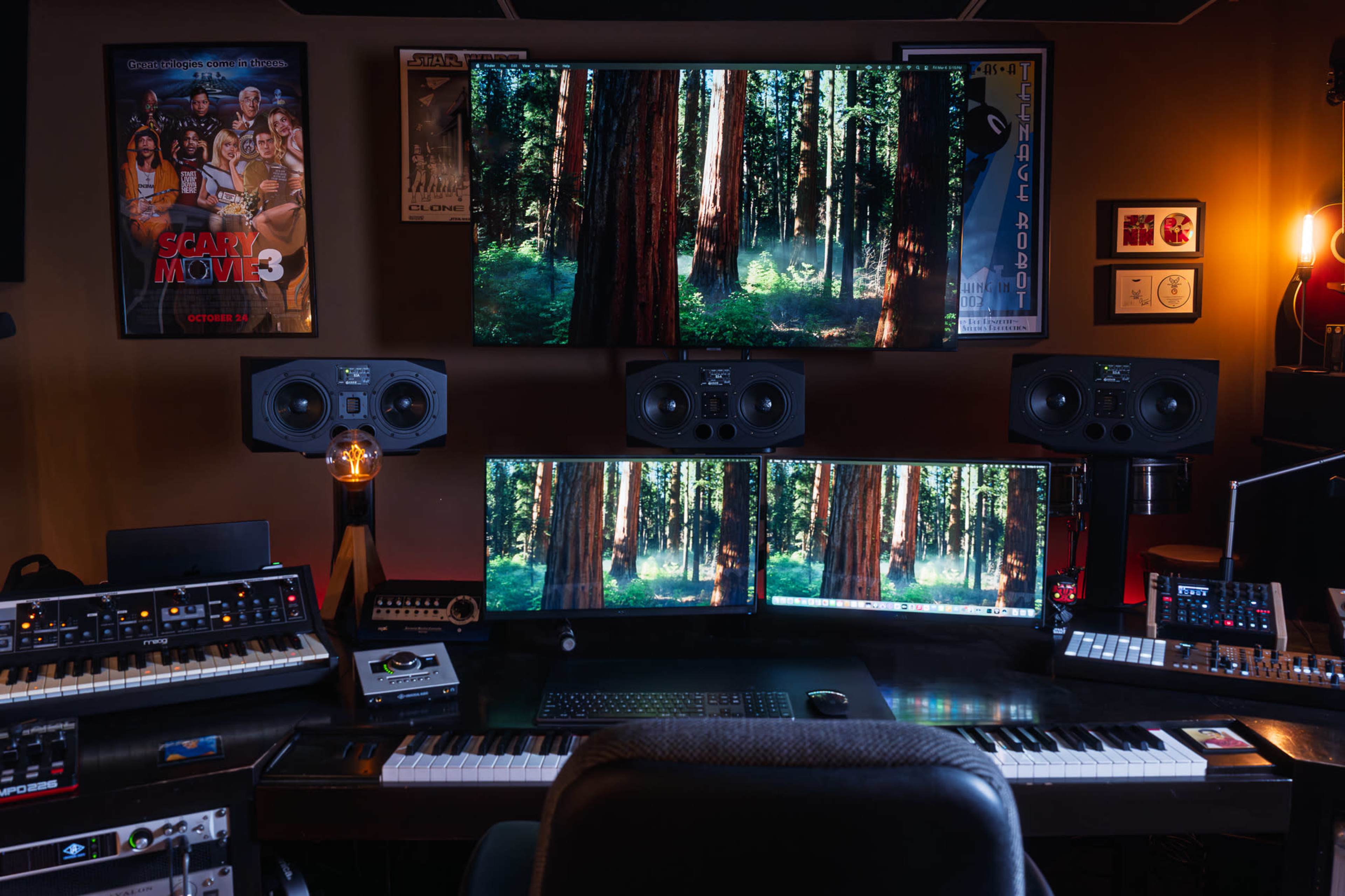 The image shows a music production setup featuring multiple monitors displaying a forest scene, studio speakers, and various electronic musical instruments arranged on a desk.