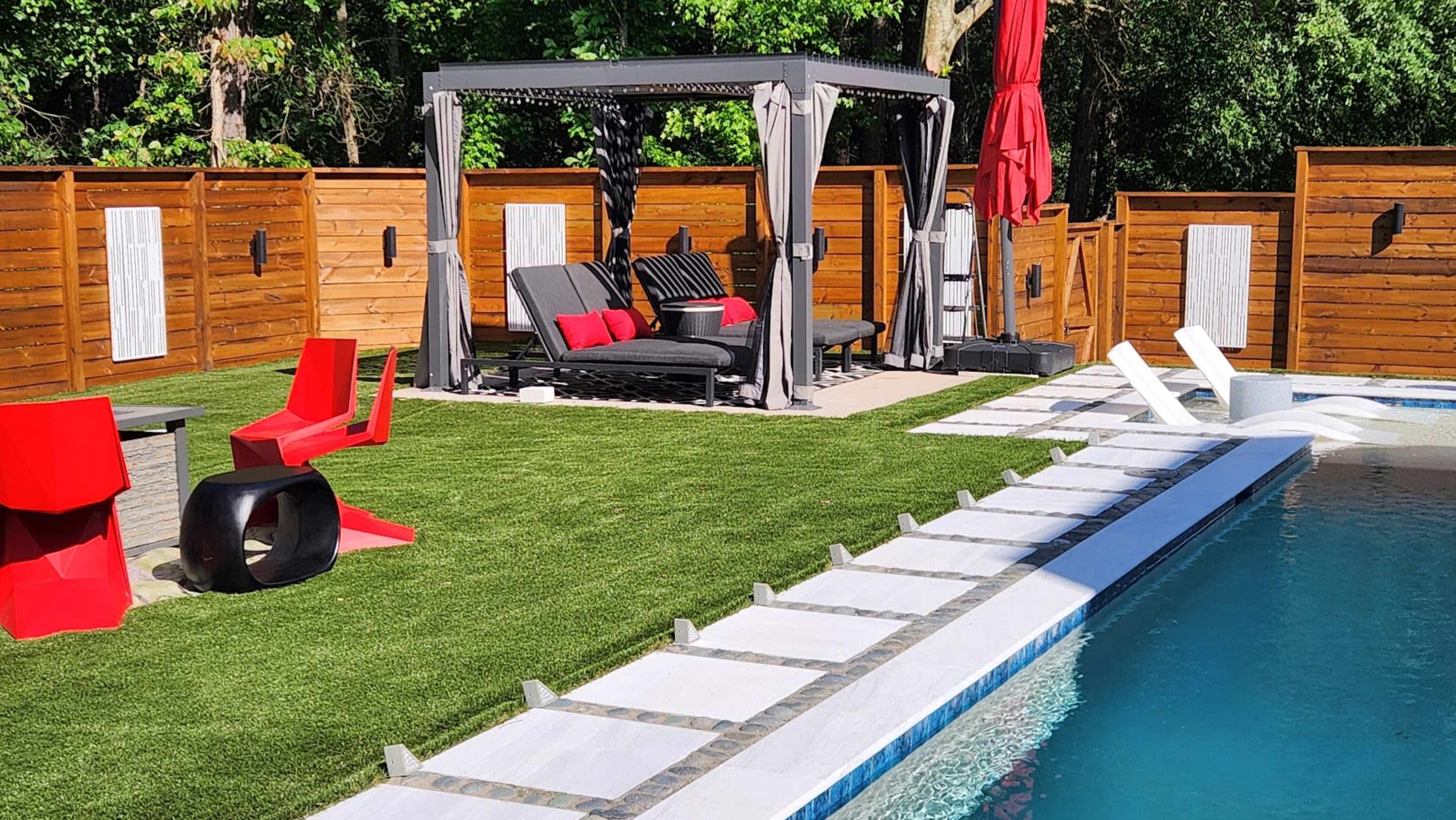 The image shows a backyard scene featuring a swimming pool, a seating area under a canopy with black furniture and red accents, and a pathway of white stones across artificial grass.