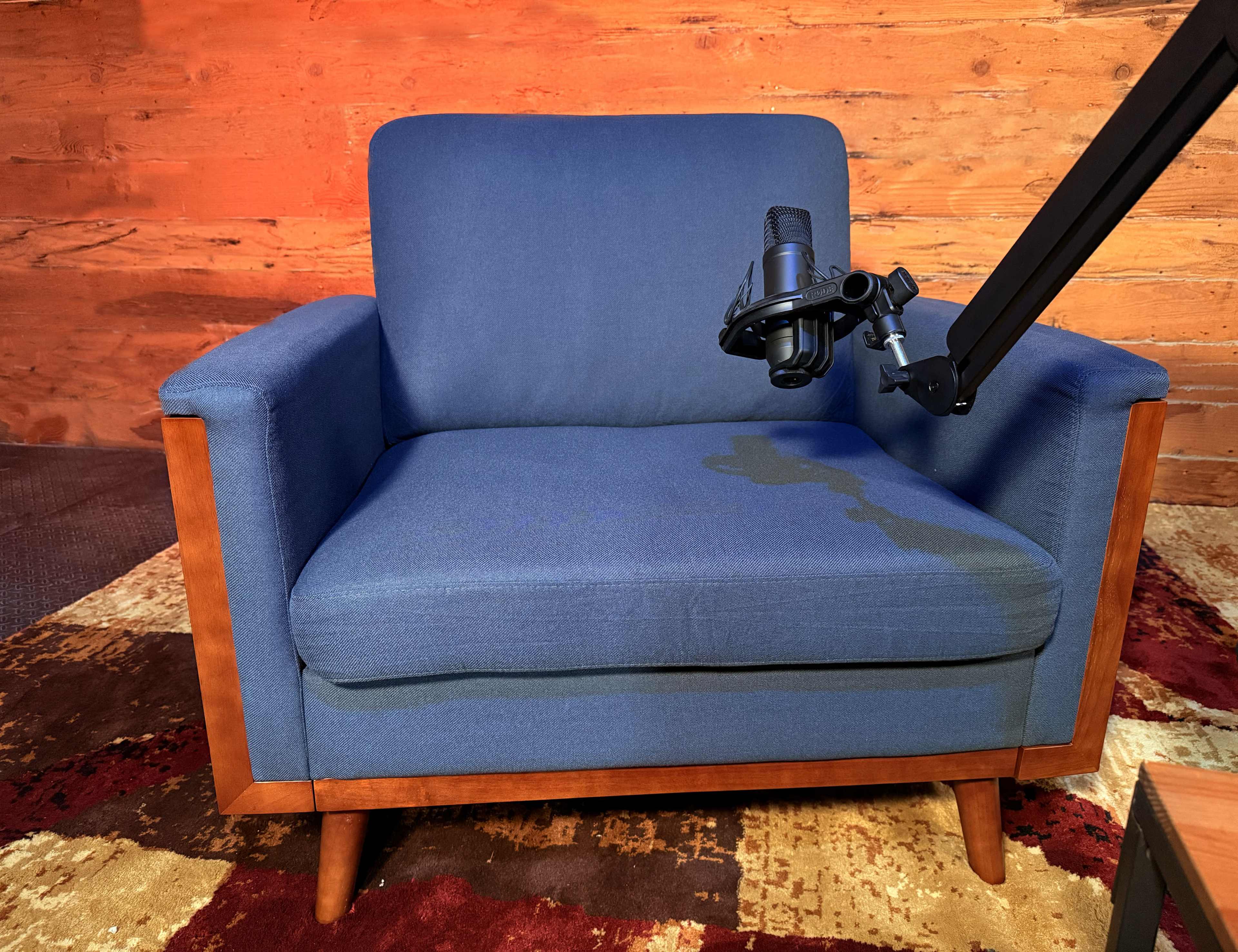 The image shows a blue armchair with wooden legs and a microphone mounted on an adjustable arm, set against a wooden wall background.