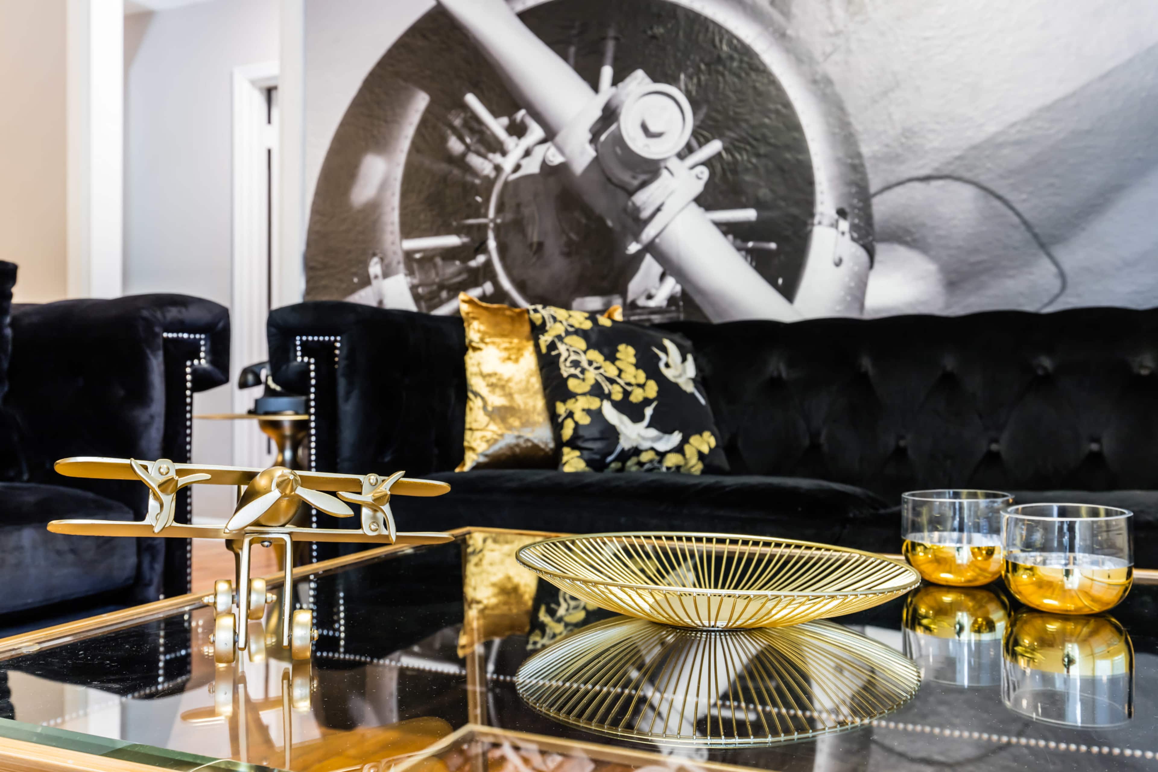 A decorative coffee table features a gold model airplane, a wire fruit bowl, and two glass candle holders, while a large black and white mural of an aircraft is displayed in the background.