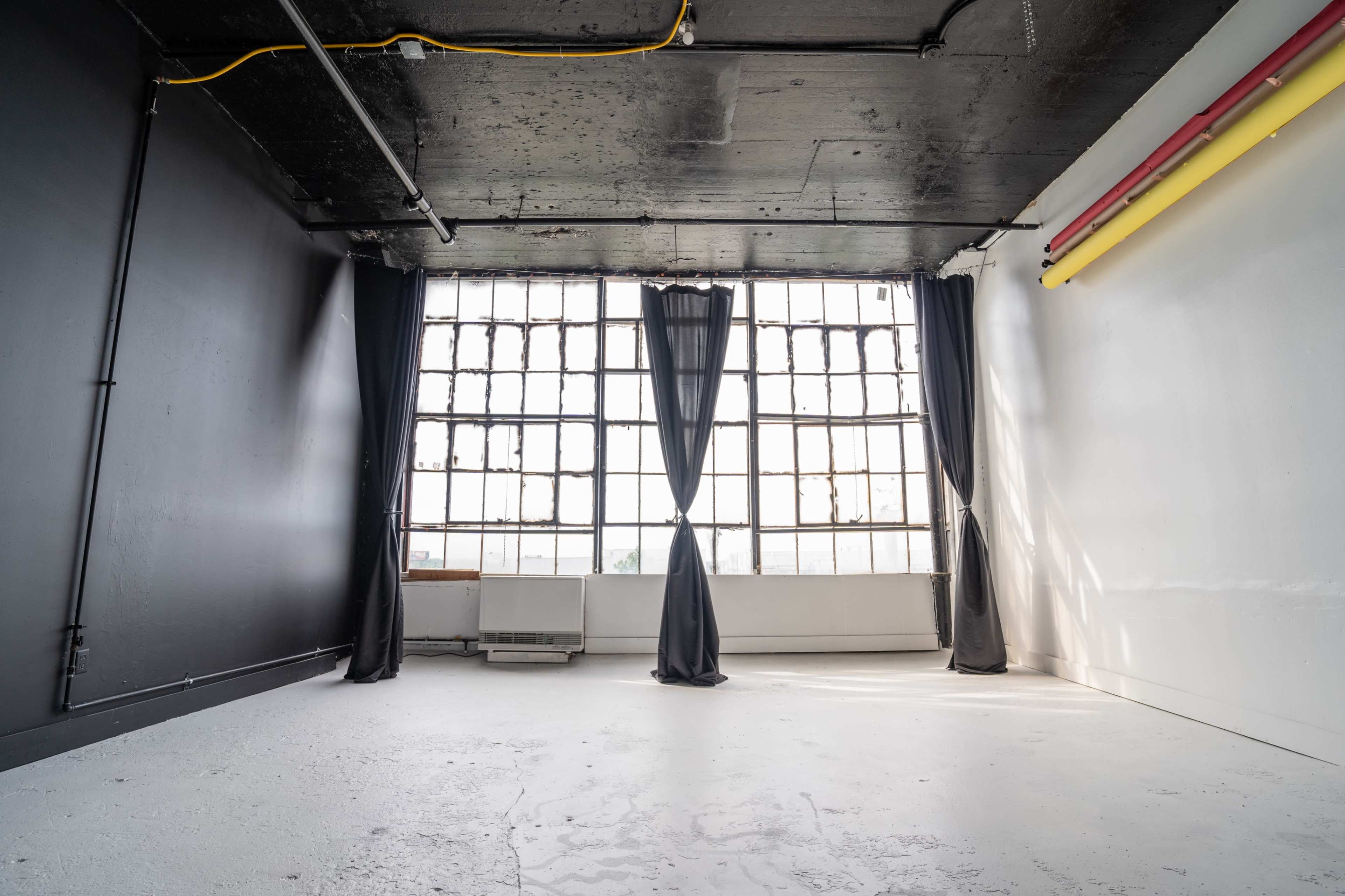 A spacious room with a concrete floor and large windows covered by black curtains, set against a backdrop of black and white walls.