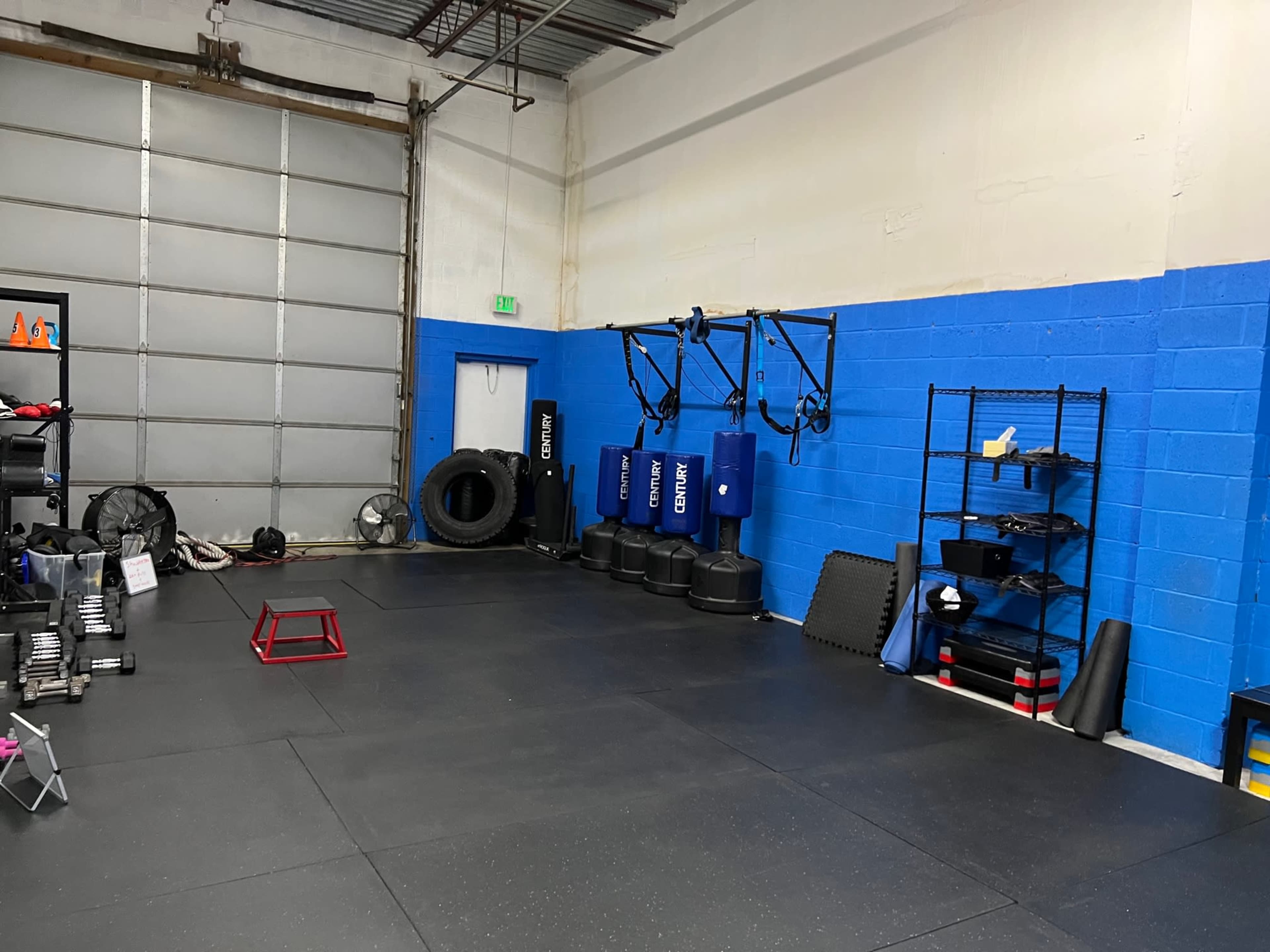 A gym space with exercise equipment including weights, a red training box, and a wall-mounted suspension trainer against a blue wall.