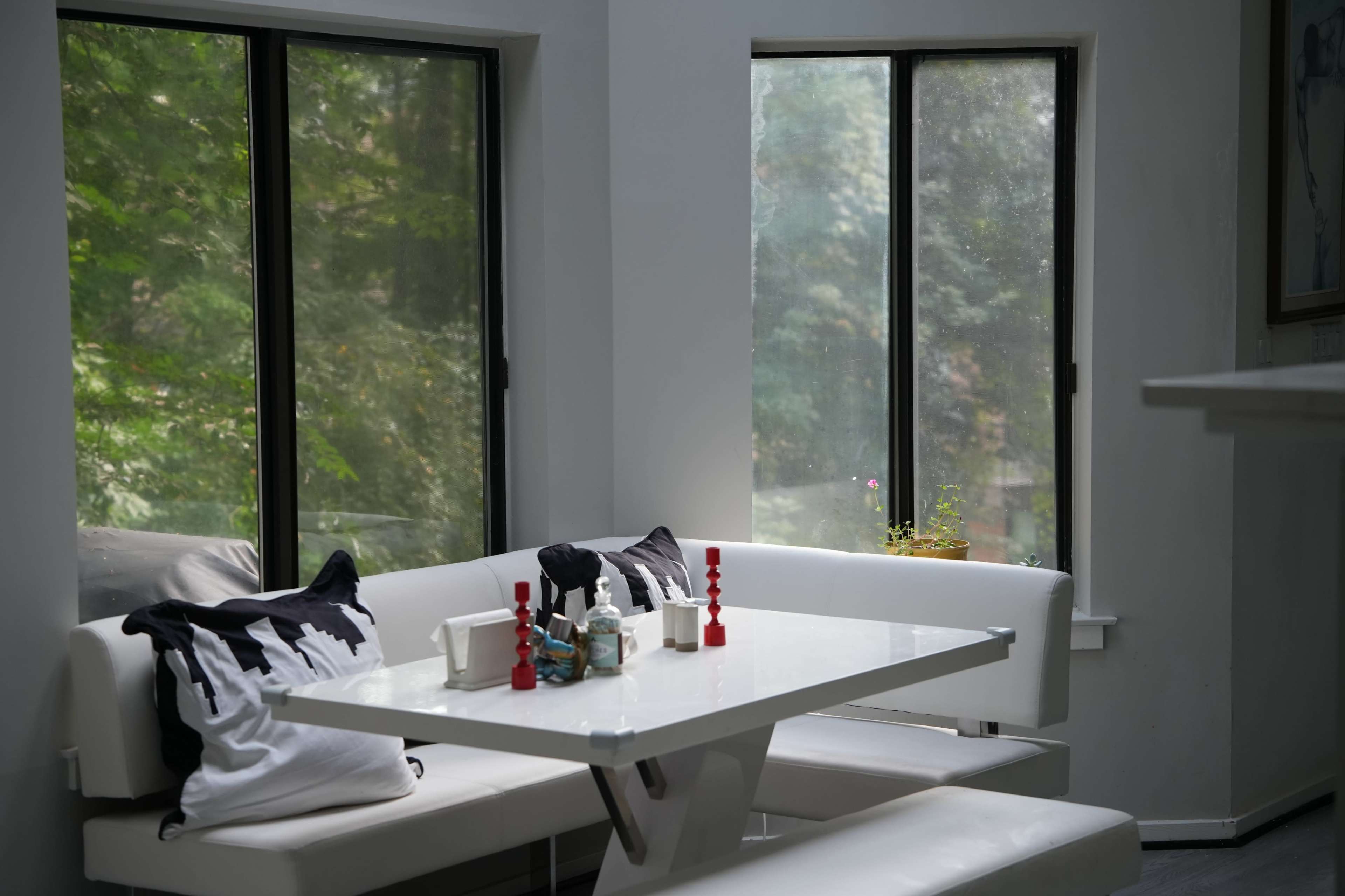 A modern dining area features a white table with upholstered seating, decorated with black and white pillows and small decorative items, alongside large windows that offer a view of greenery outside.