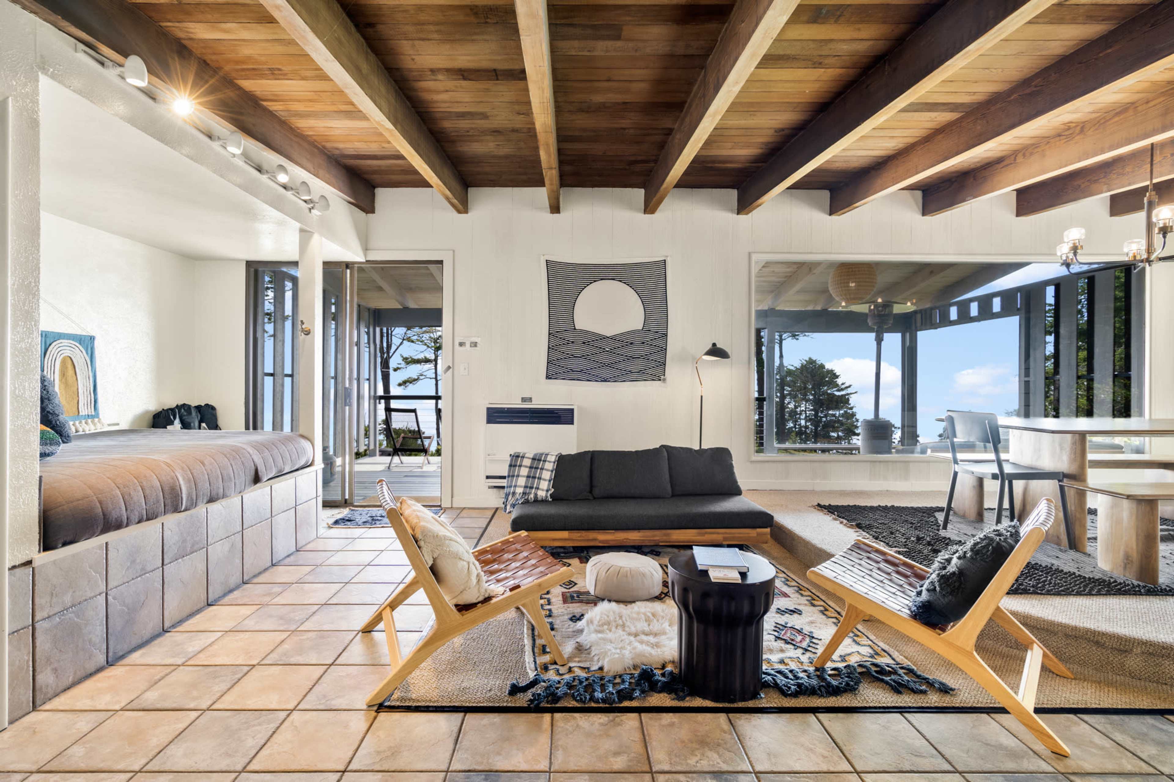 The image shows a modern living space with wooden beams, a sectional sofa, and large windows that offer a view of the outdoors.