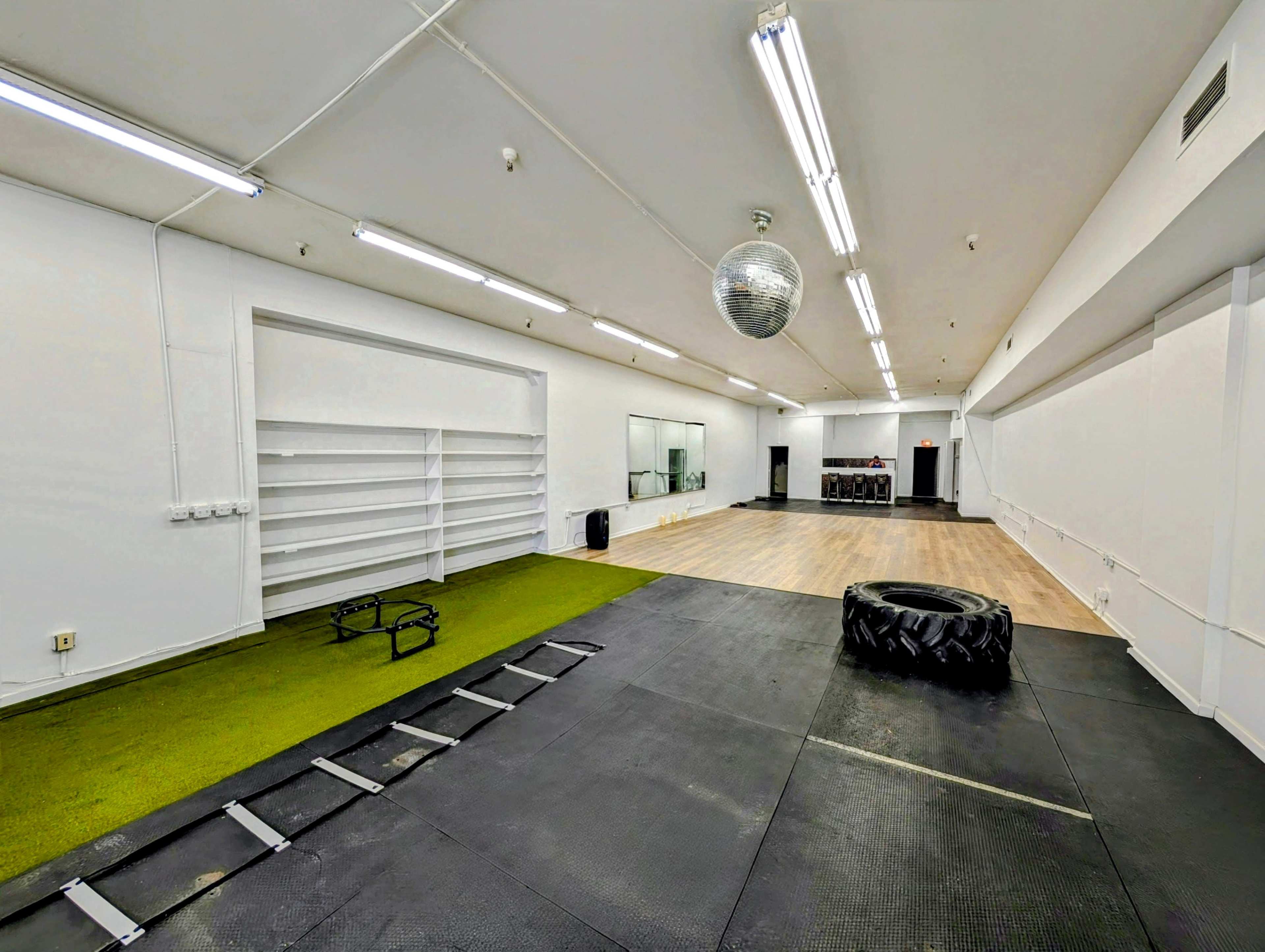 The image shows a spacious workout area with a green exercise mat, a large tire, a disco ball overhead, and wooden flooring on one side.