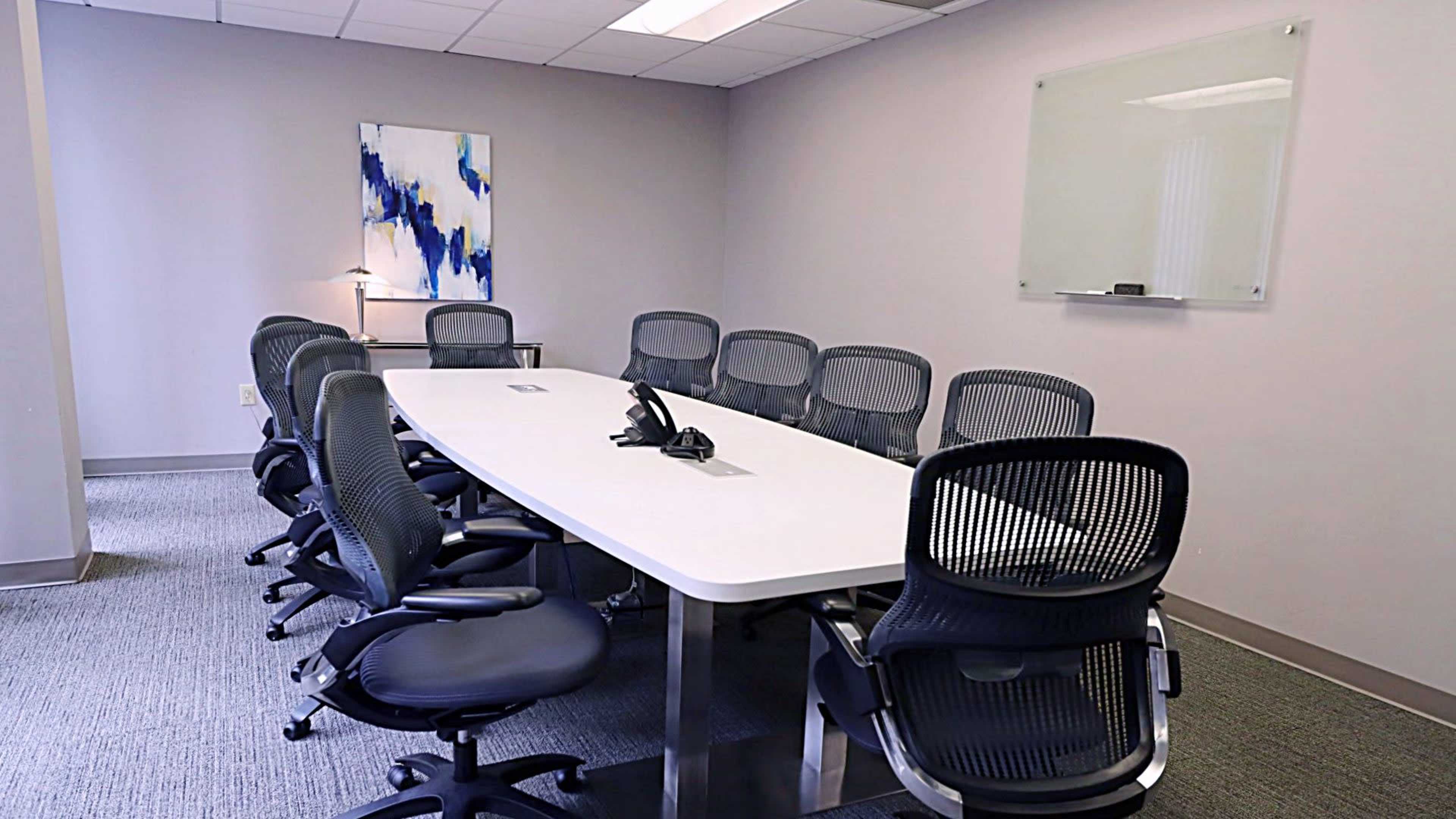 The image shows a modern conference room with a long white table, several ergonomic chairs, and a wall-mounted whiteboard.