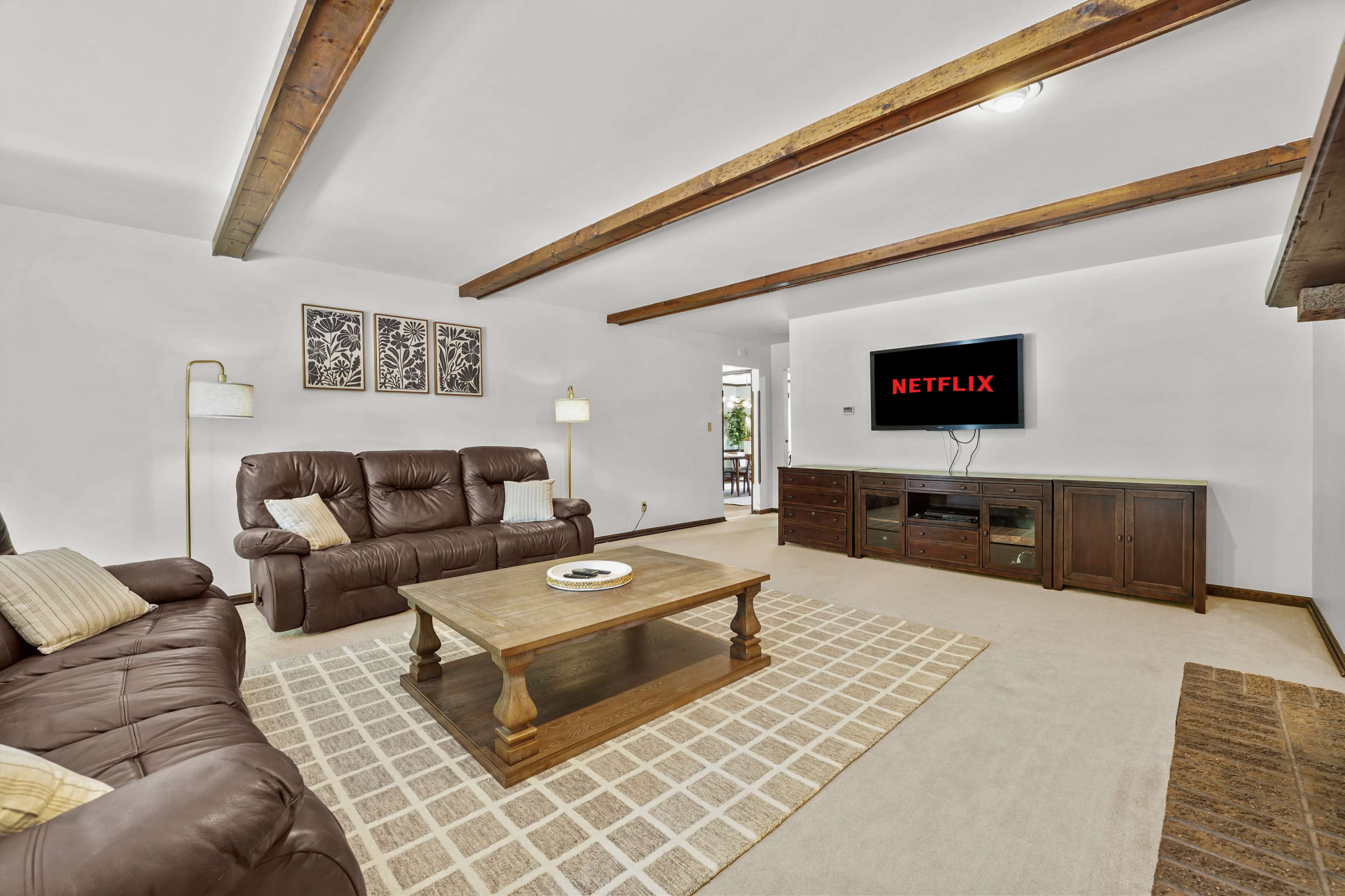 The living room features brown leather recliners, a wooden coffee table, and a television displaying the Netflix logo, with exposed wooden beams on the ceiling.
