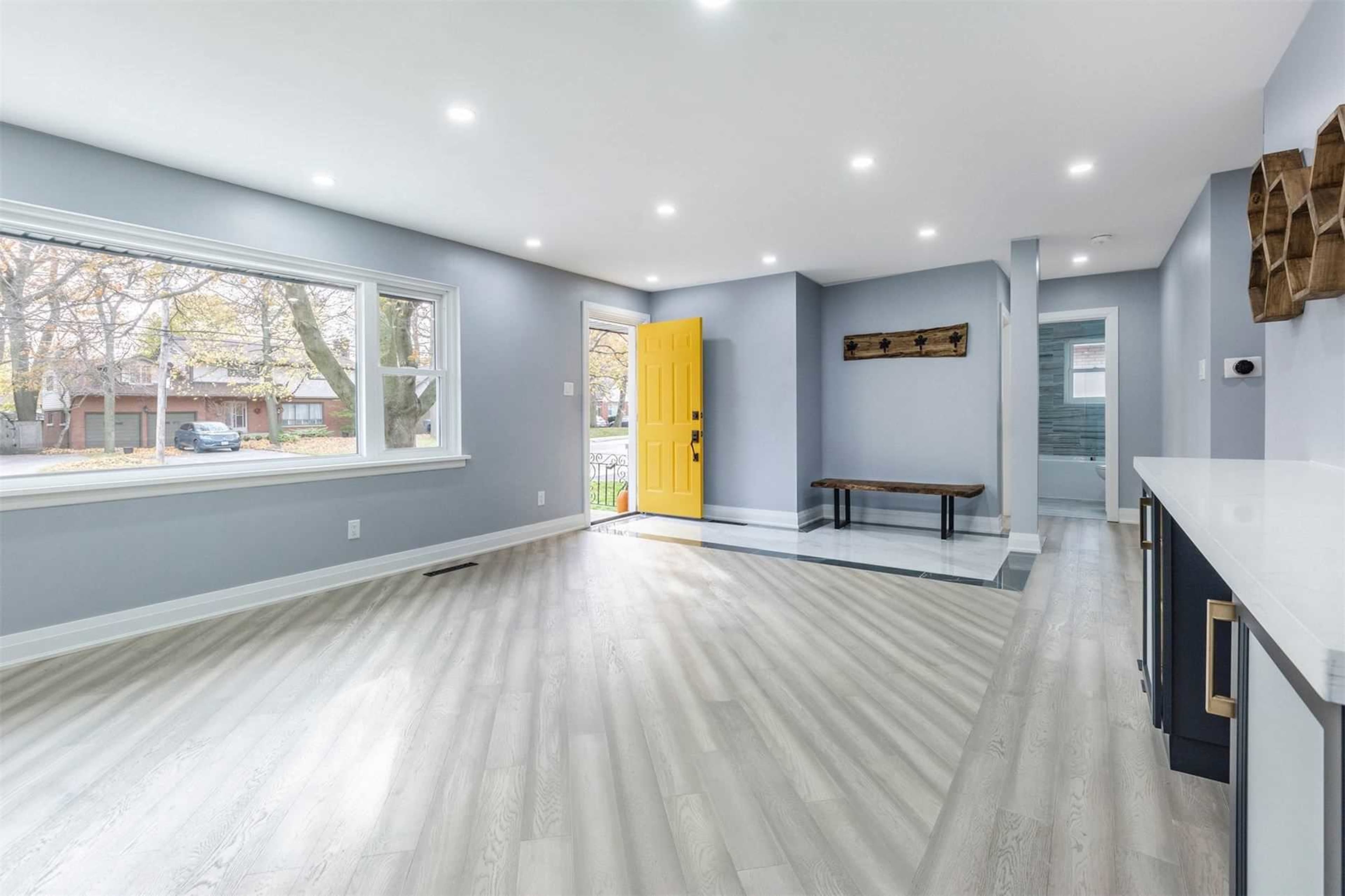 The image shows a spacious, well-lit interior room with light gray walls, a yellow front door, and a wooden bench against one wall.