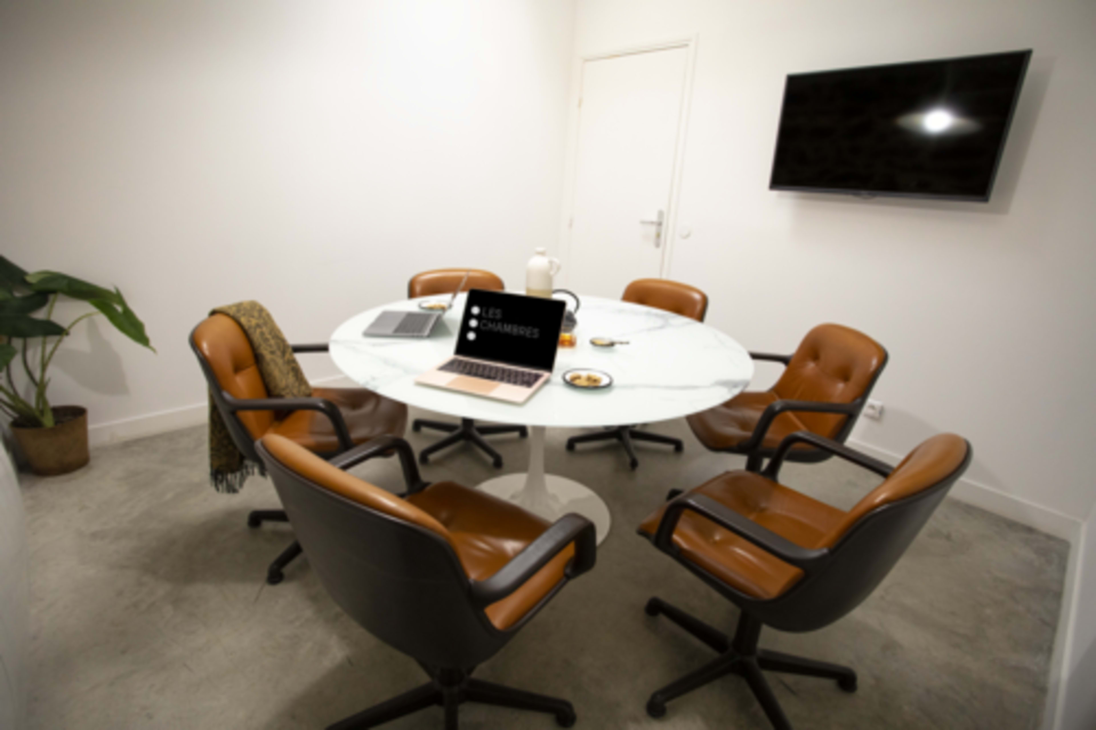 A modern conference room features a round marble table surrounded by six brown leather chairs, with a laptop and a teapot on the table, and a television on the wall.