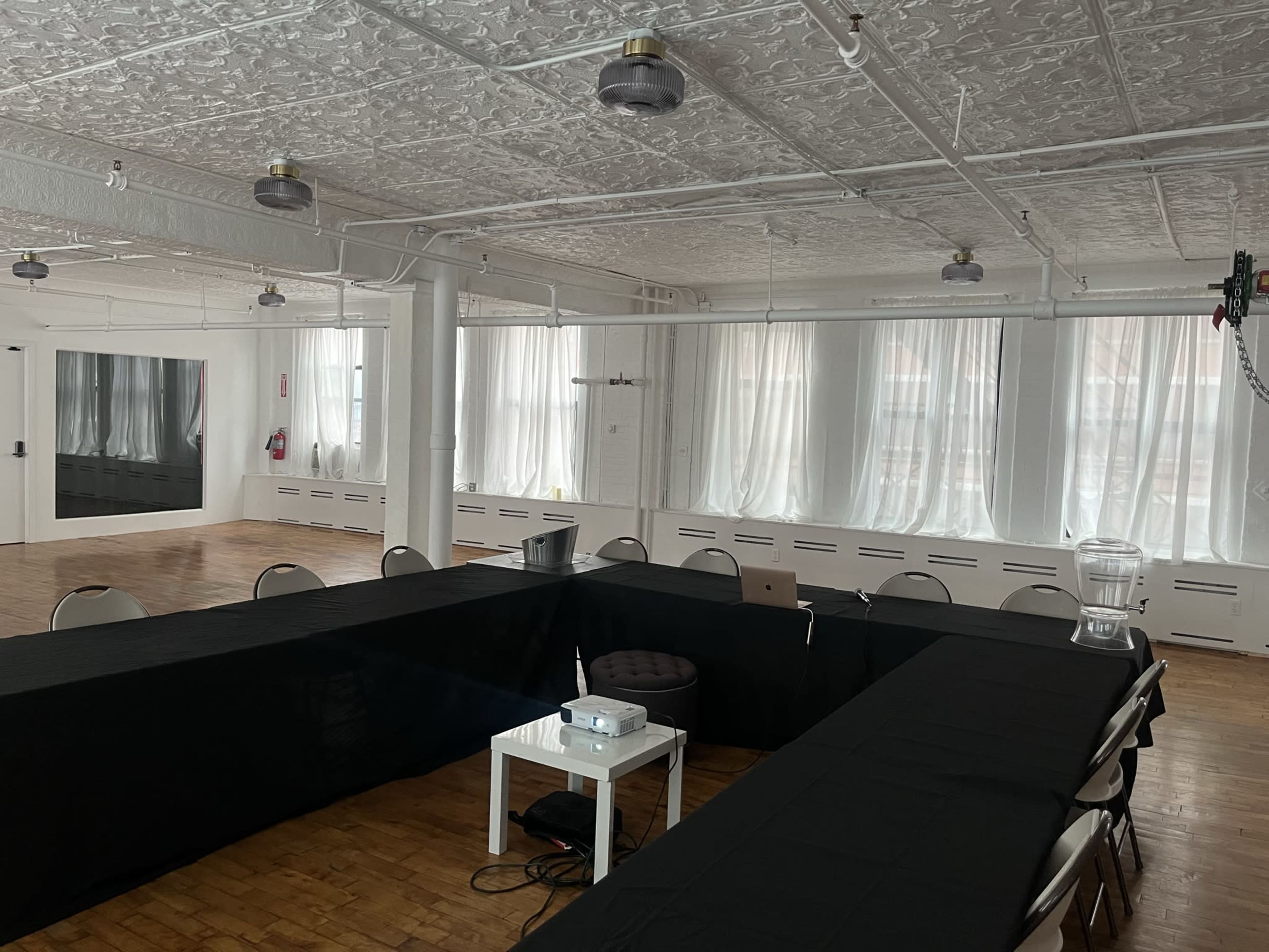 A rectangular meeting room features a setup of black tables arranged in a U-shape, with chairs around them and large windows letting in natural light.