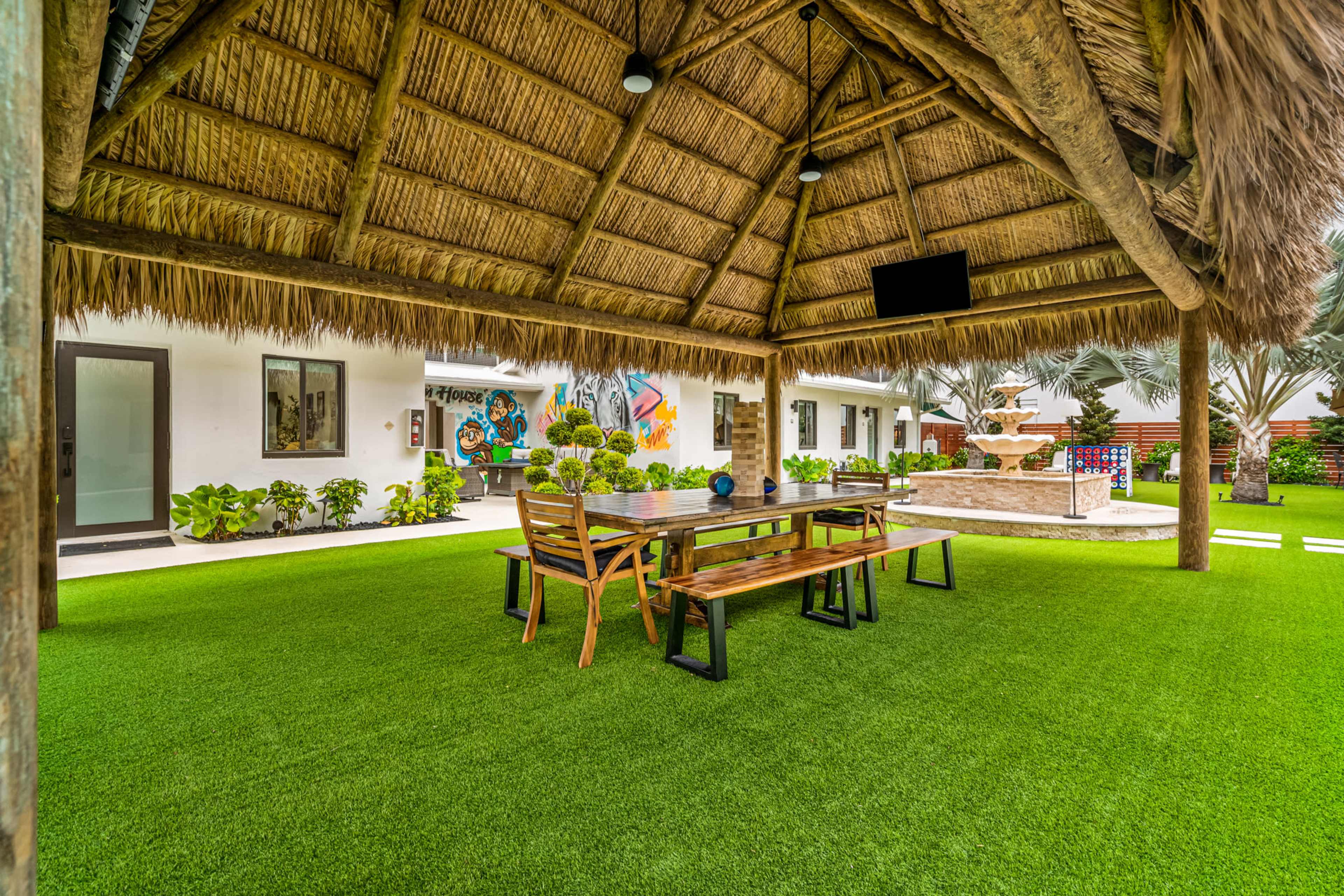 A thatched-roof pavilion with wooden tables and chairs is surrounded by green grass and tropical plants, with buildings and artwork visible in the background.