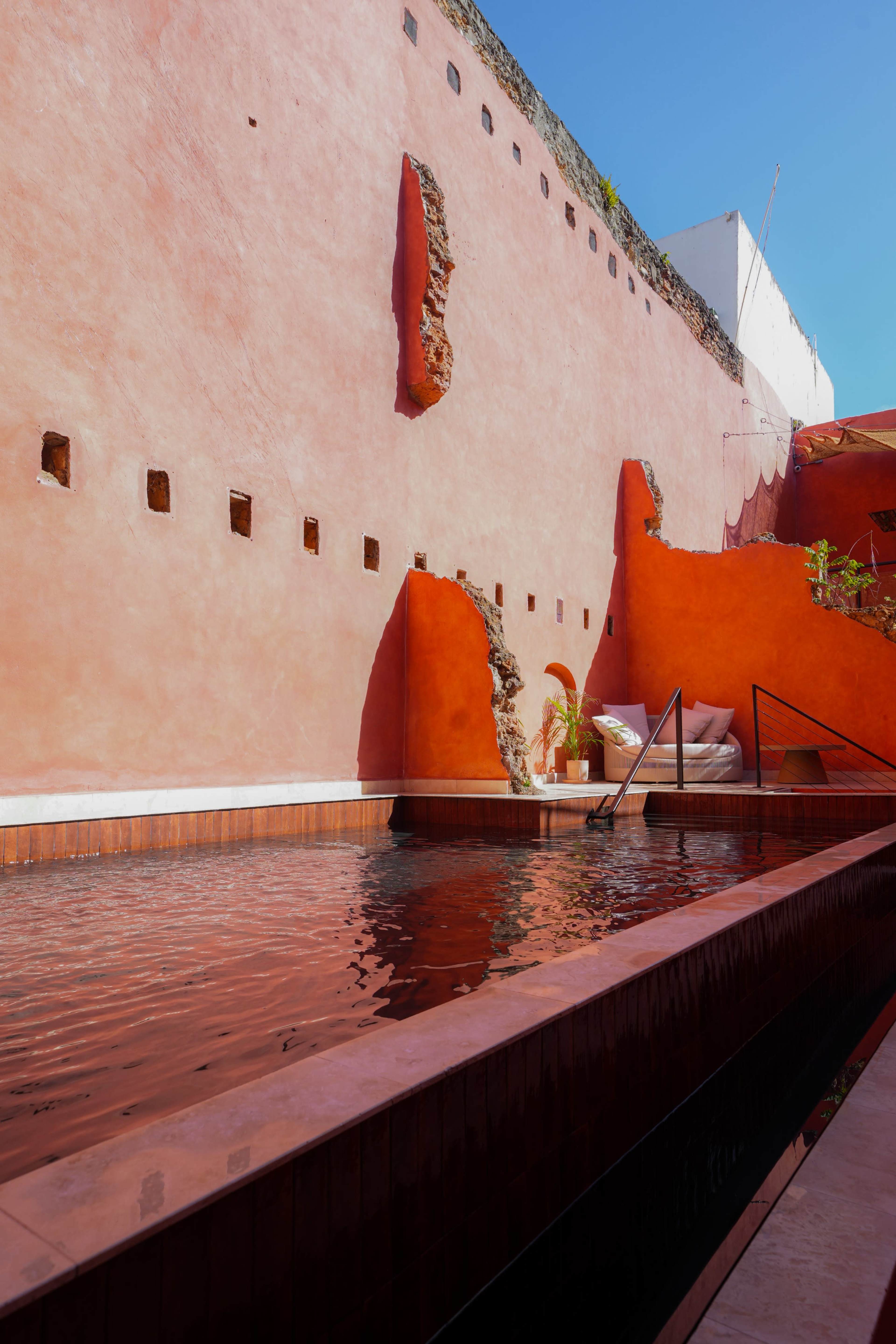 The image depicts a tranquil outdoor pool area surrounded by vibrant orange walls and architectural elements, with clear blue skies overhead.
