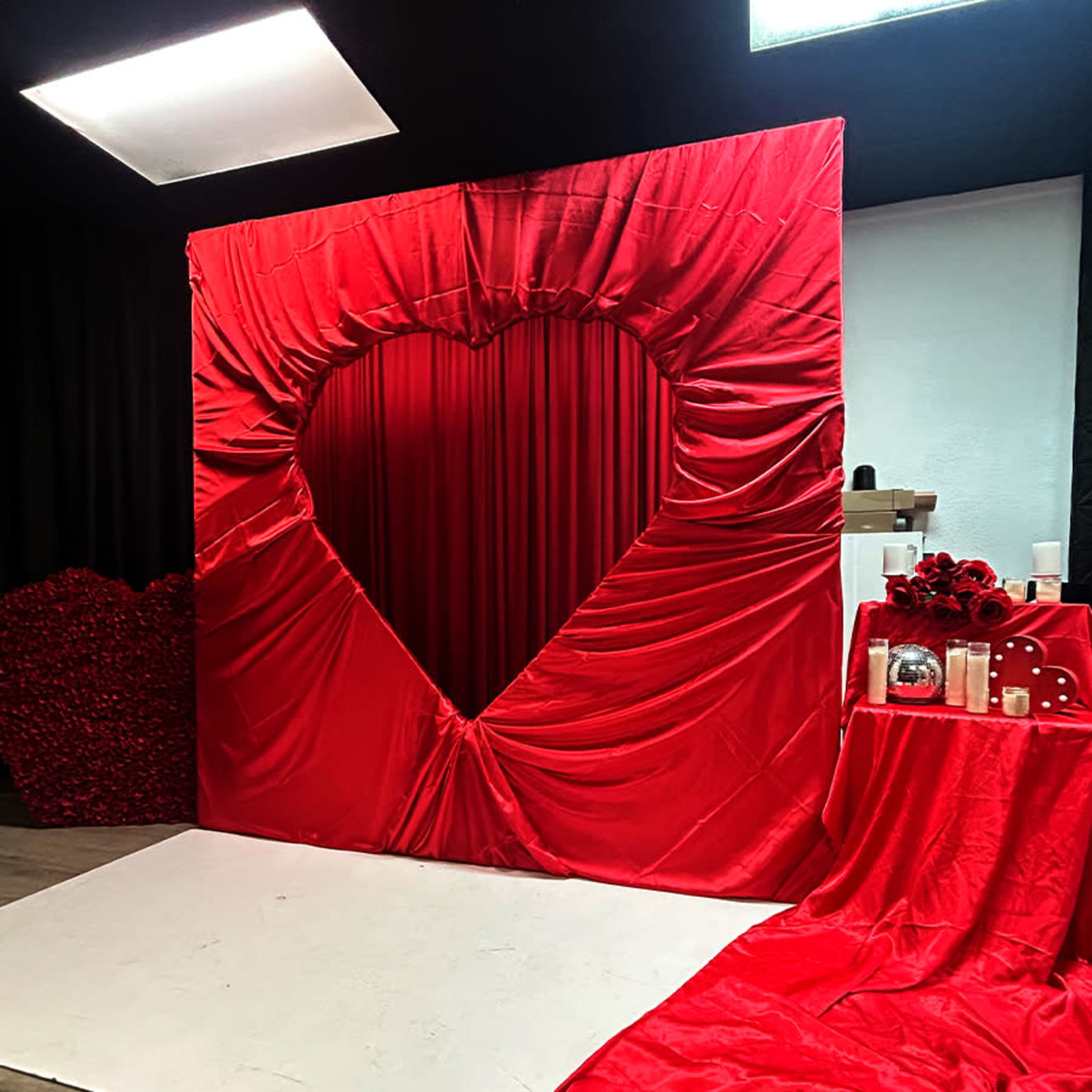 A large red fabric backdrop shaped like a heart is displayed in a studio setting, accompanied by decorative elements such as candles and roses.