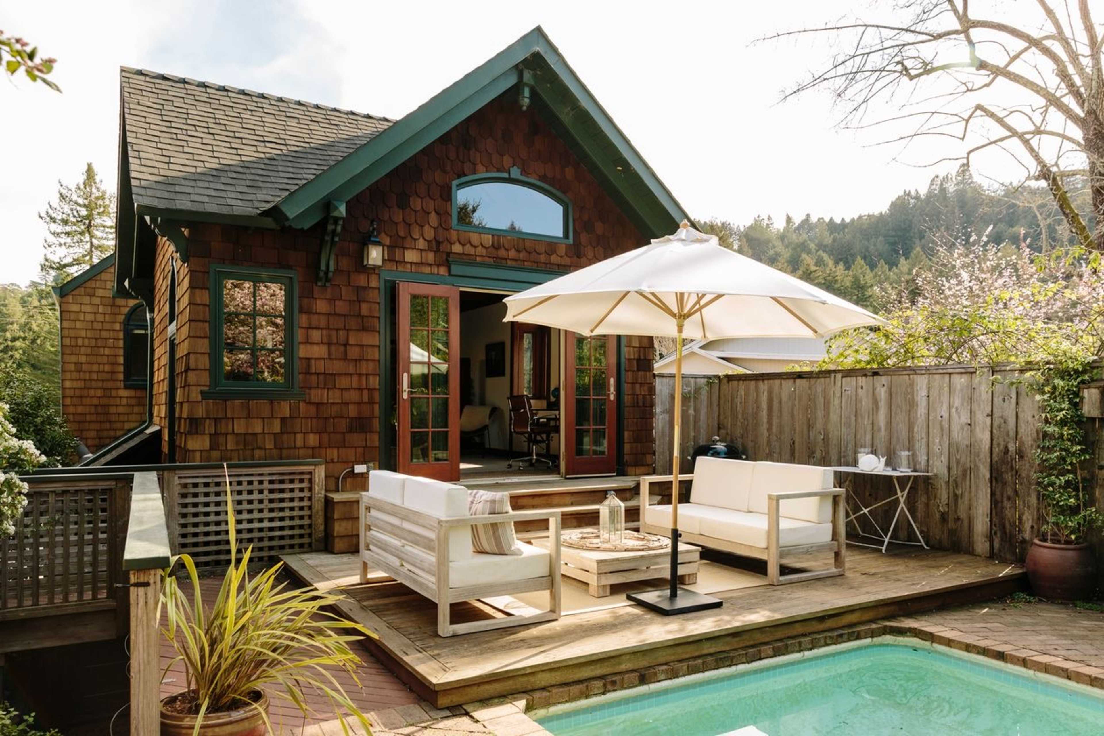 A wooden deck features modern outdoor furniture and an umbrella beside a swimming pool, adjacent to a cottage with stone siding and large windows.